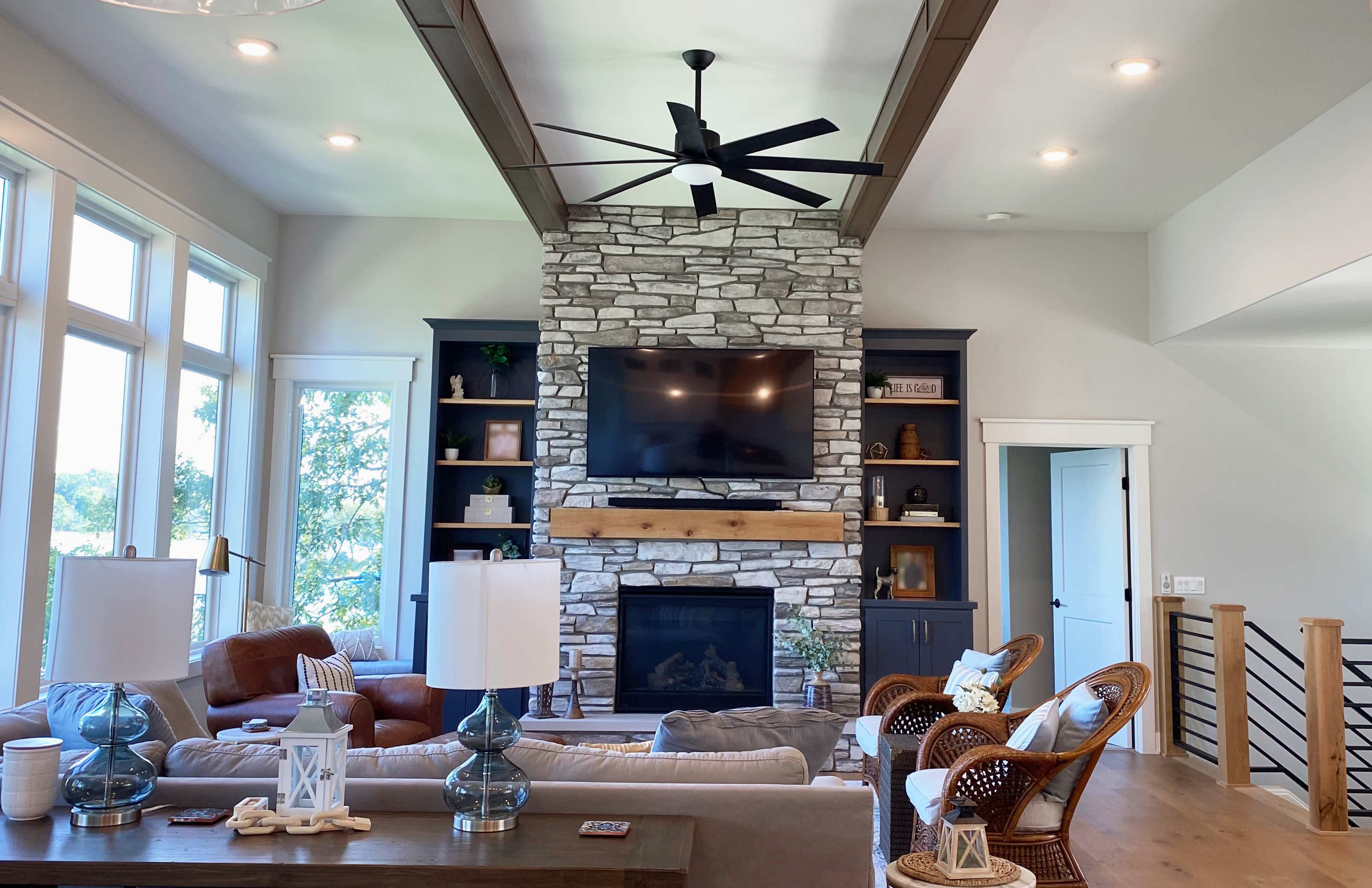 The living room features a stone fireplace with a mounted television above, surrounded by built-in shelves, wooden beams, and large windows allowing natural light to enter.