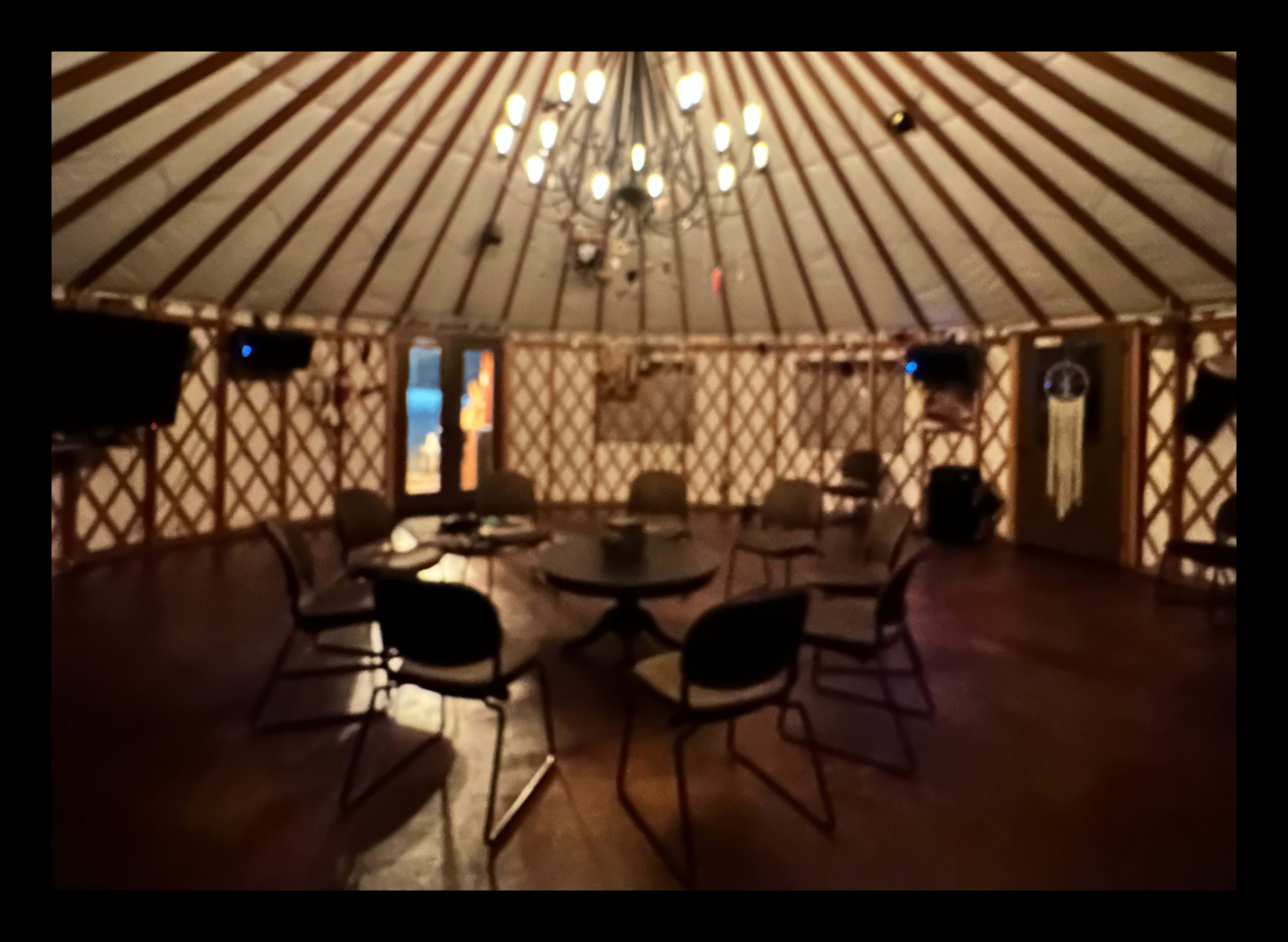 The interior of a yurt, featuring a circular layout with a central table surrounded by chairs and illuminated by a chandelier.