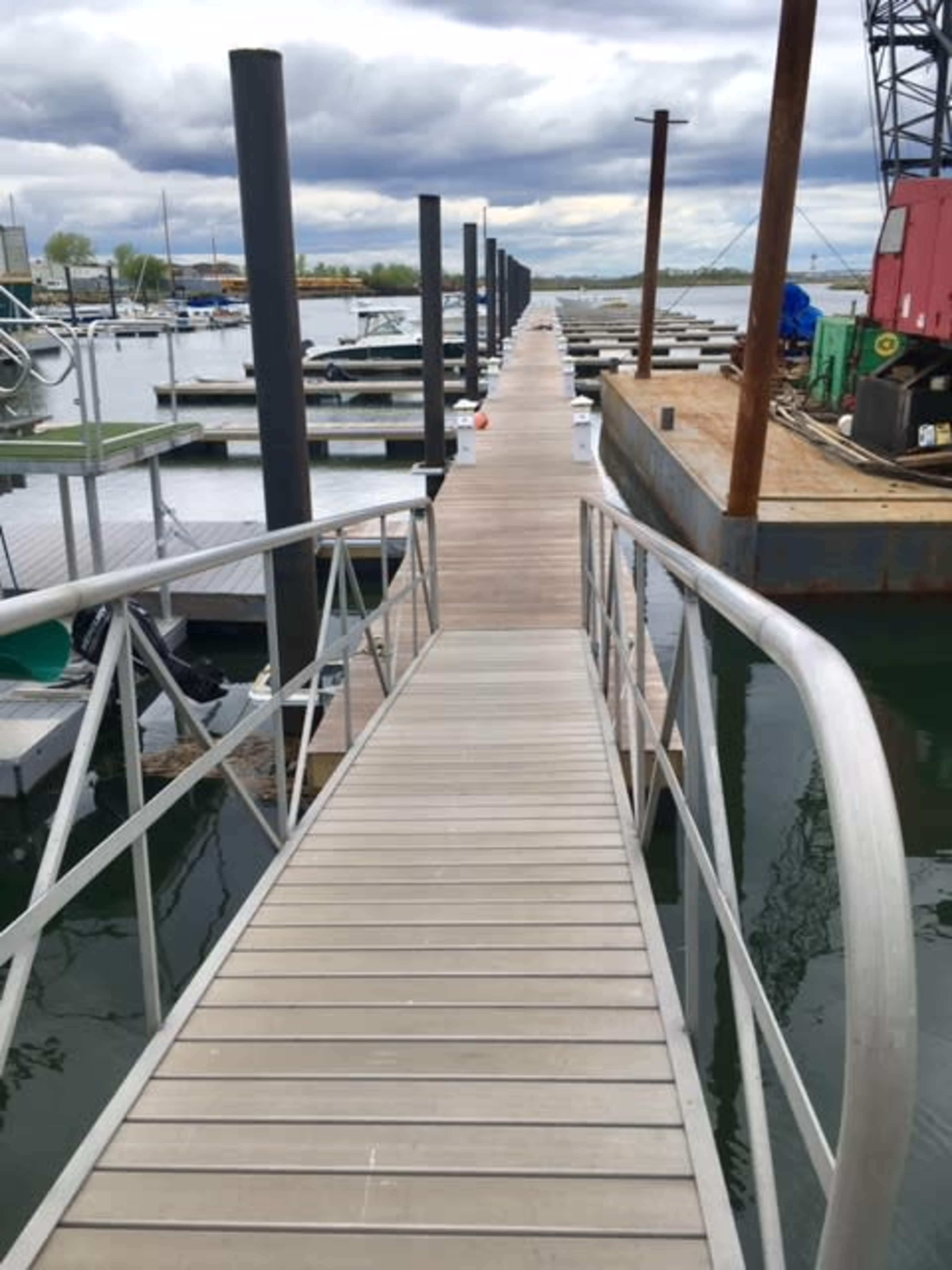 The image shows a wooden walkway leading to multiple boat slips at a marina, with metal pilings and cloudy skies above.