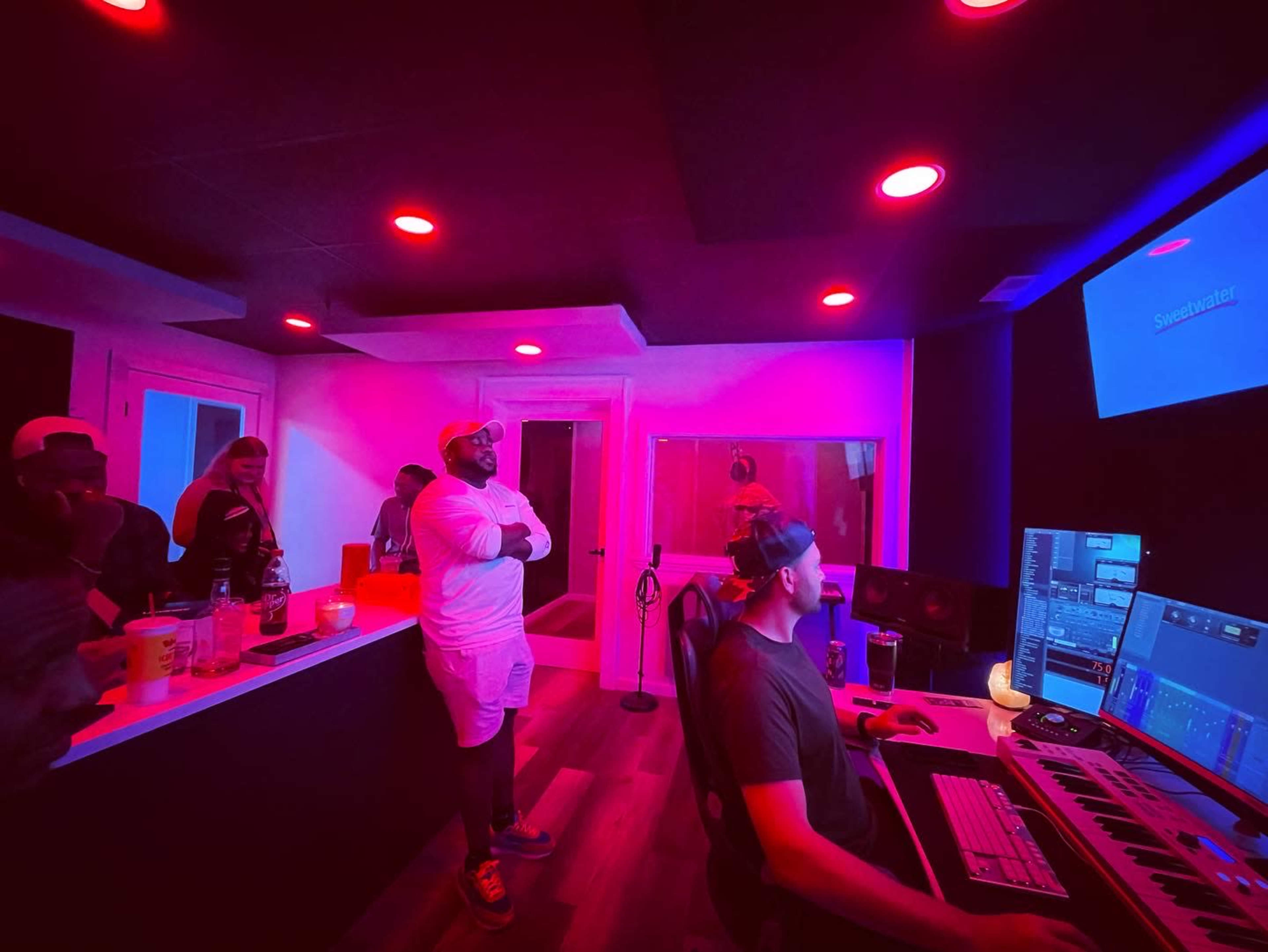 A group of people observes a music production session in a dimly lit recording studio with pink and red lighting.