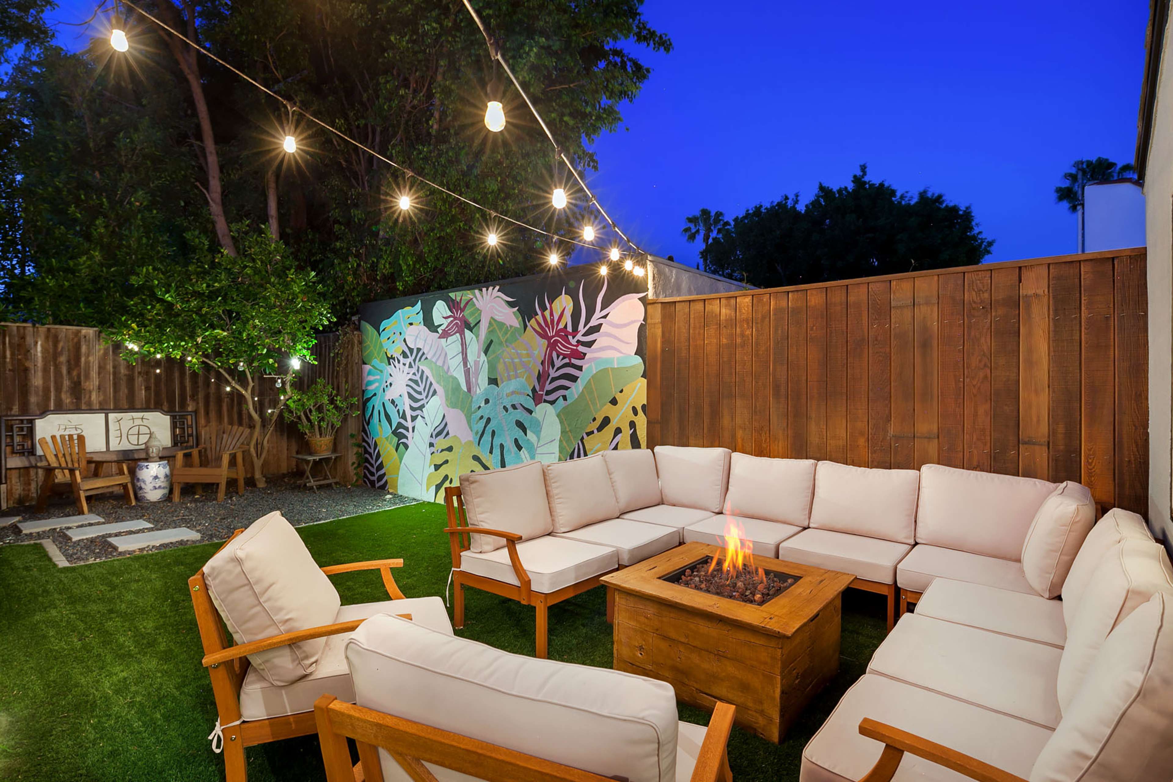 A backyard seating area features a circular arrangement of white couches around a wooden fire pit, with string lights overhead and a colorful mural on the back wall.