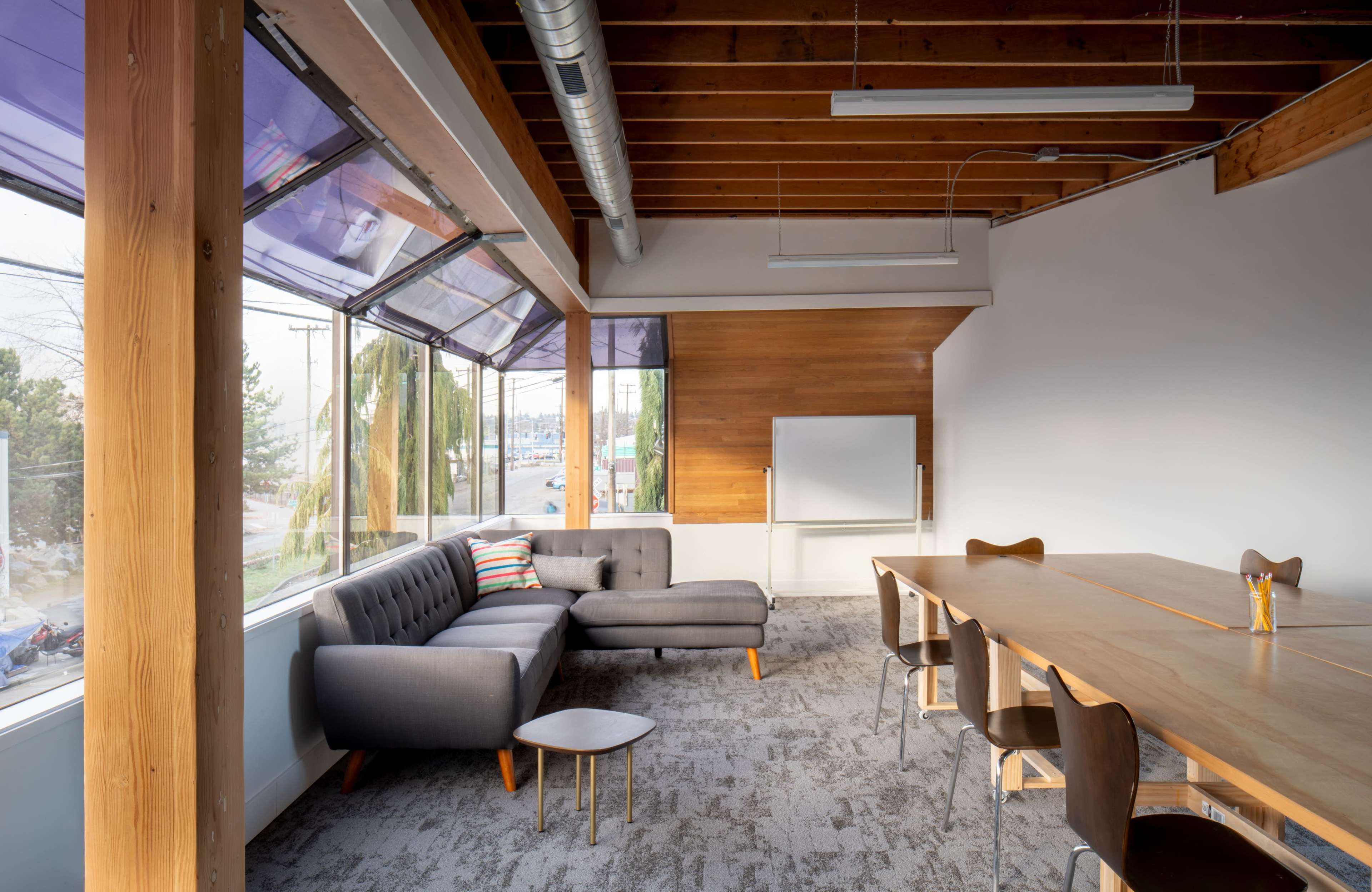 A modern office space featuring a gray sectional sofa, a wooden conference table with chairs, and large windows with natural light.
