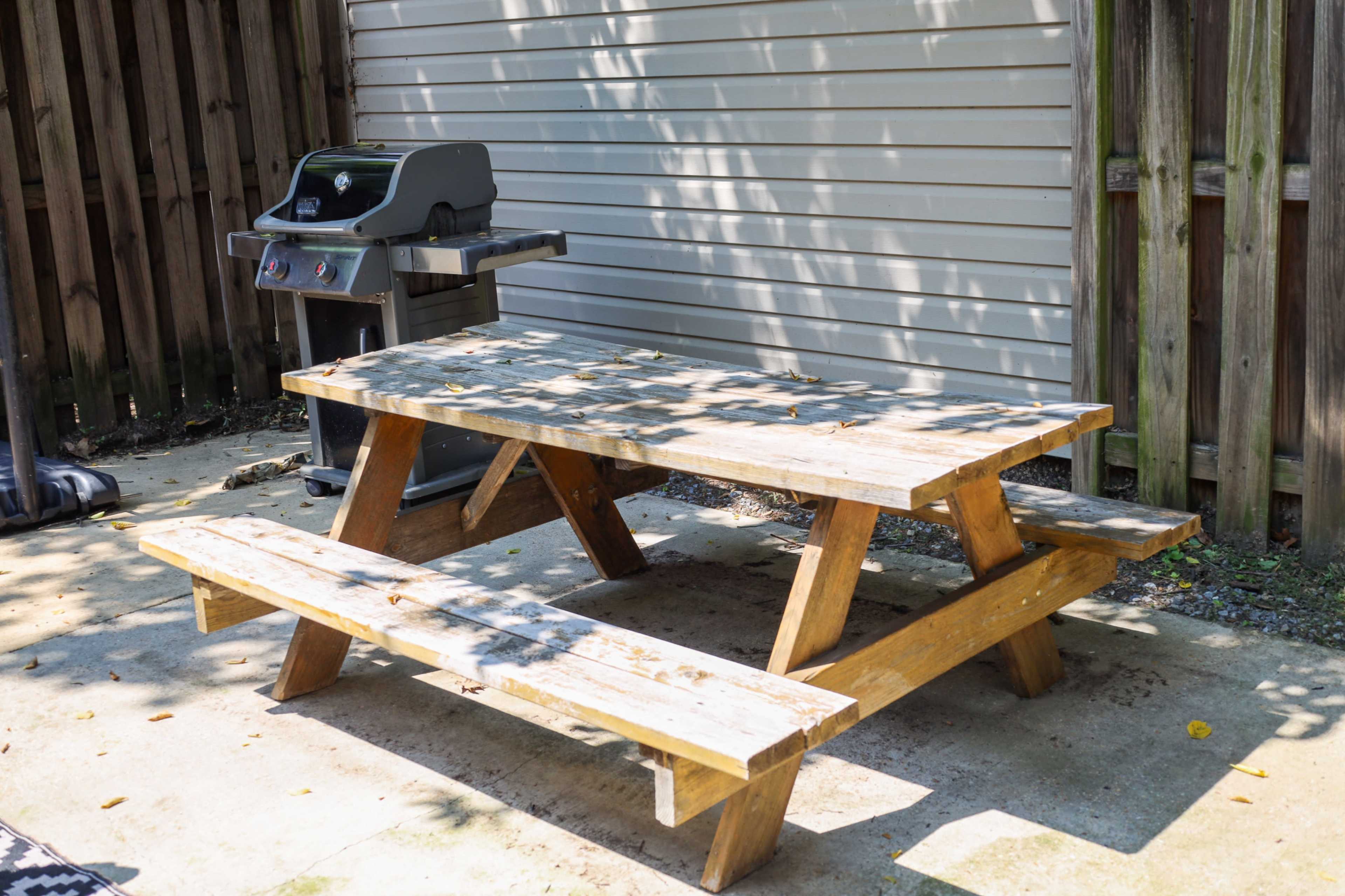A wooden picnic table is positioned next to a grill in a back patio area lined with a wooden fence.