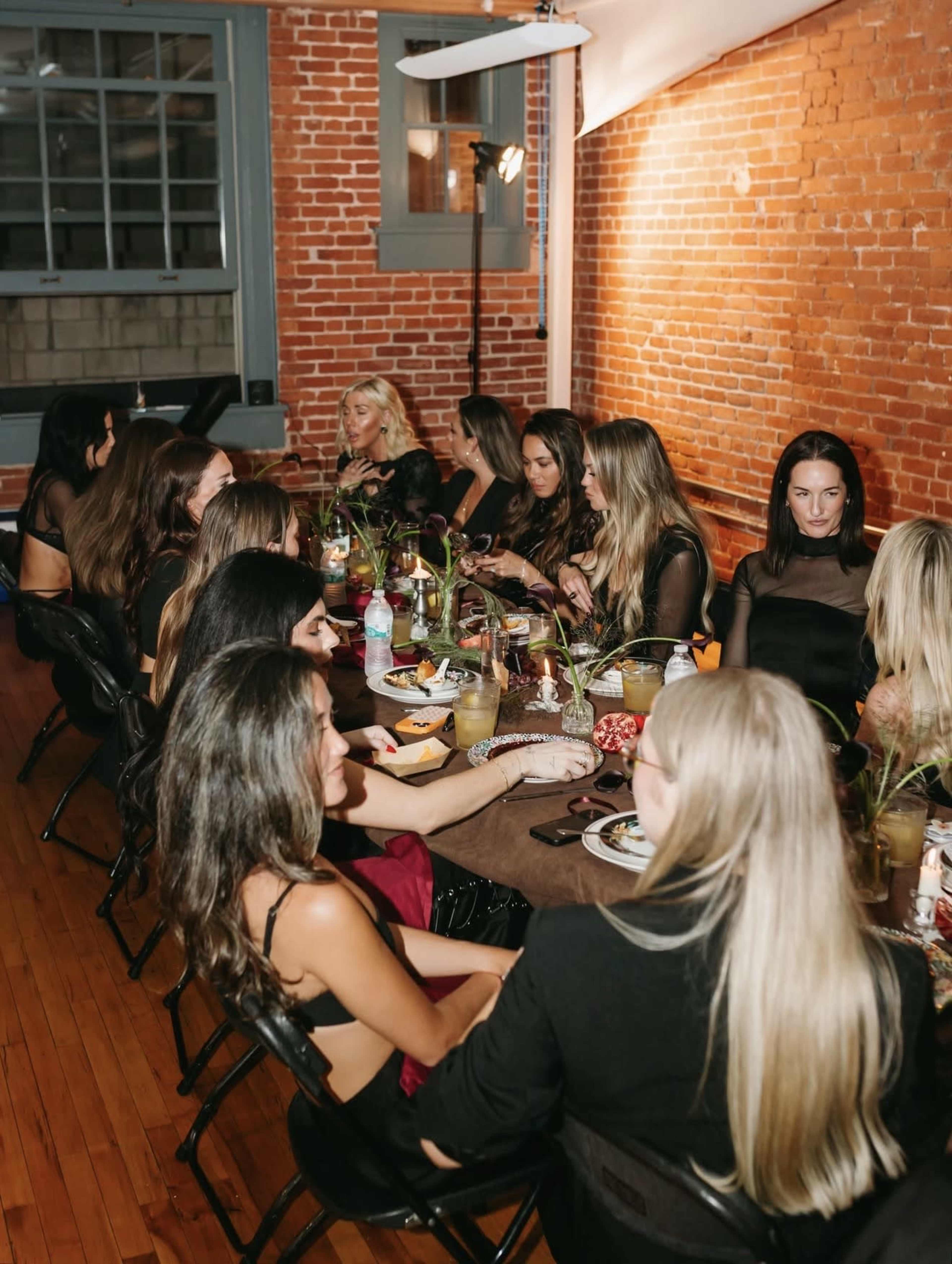 A group of women is sitting around a long table adorned with food and drinks in a brick-walled room.
