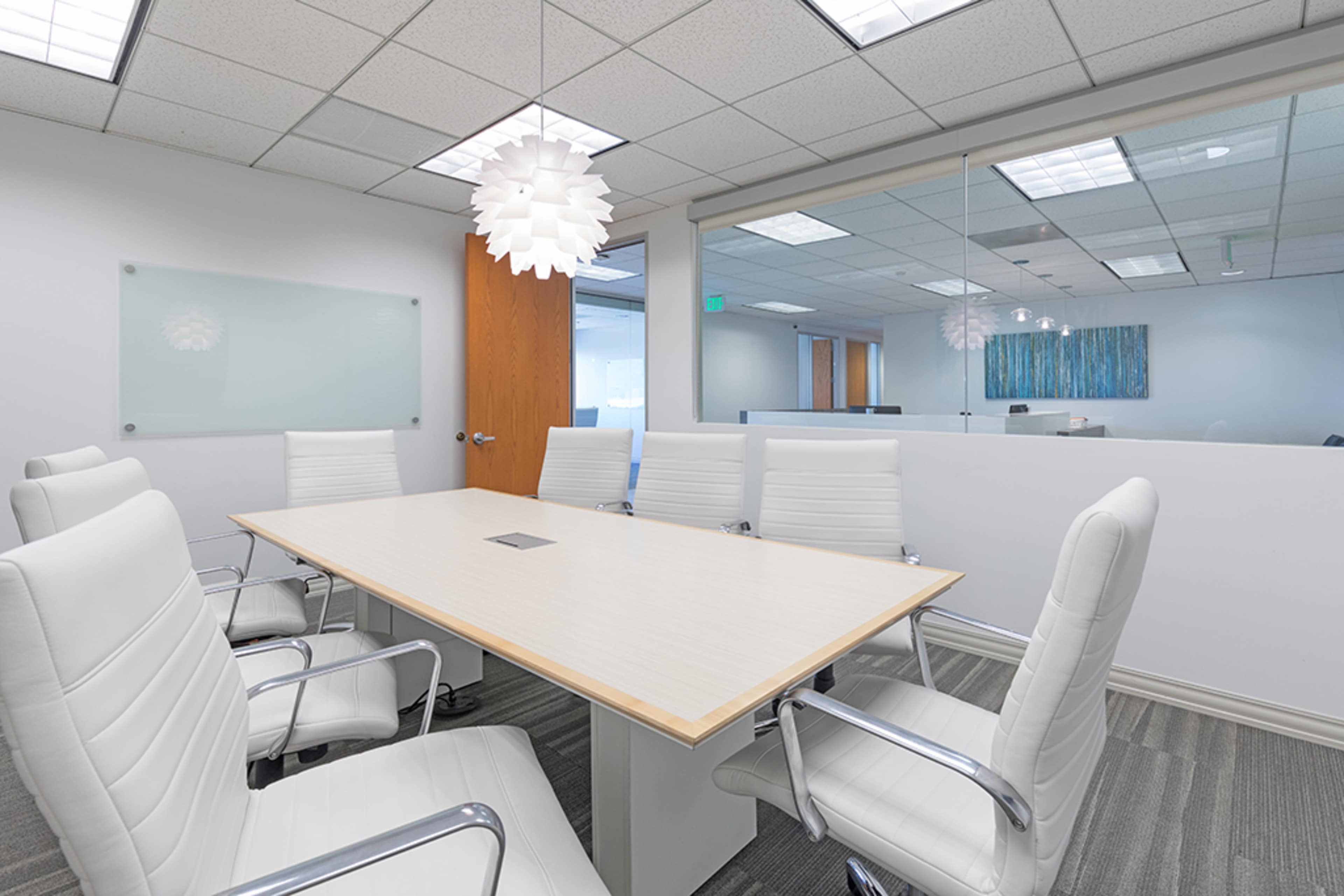 A modern conference room features a large wooden table surrounded by white chairs and a pendant light above.