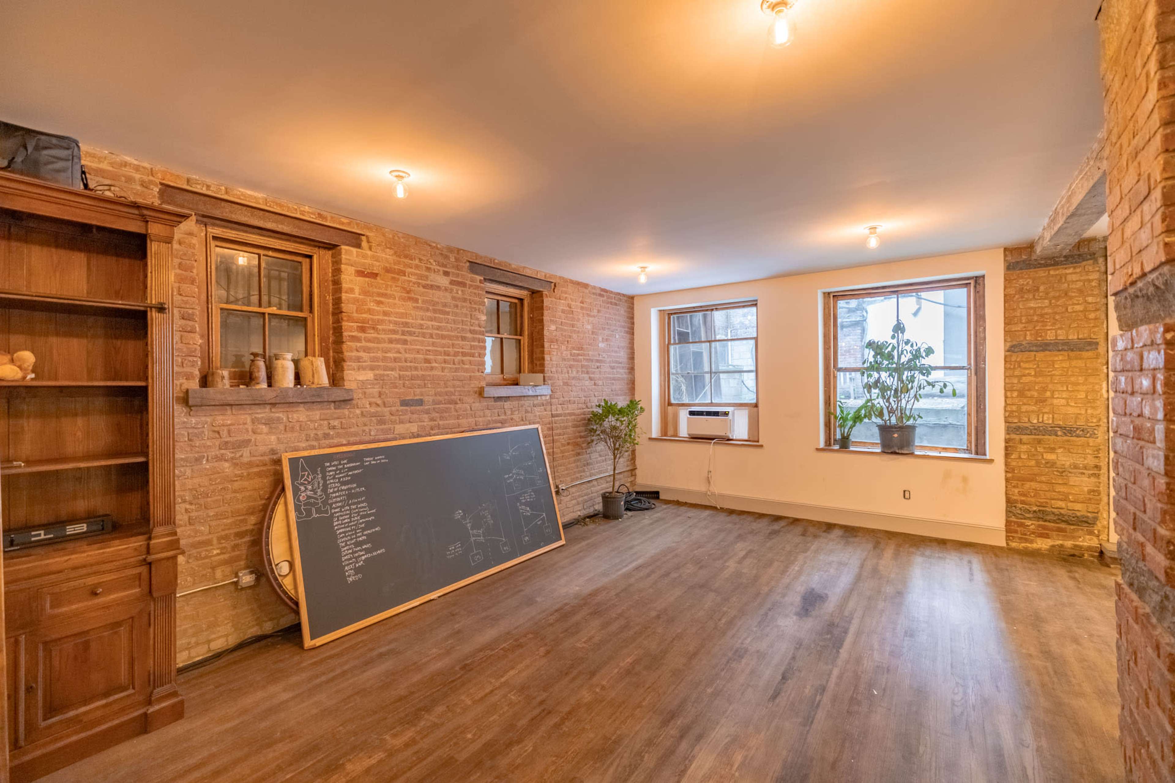 The room features exposed brick walls, hardwood floors, a large chalkboard, and two windows with plants on the sill.
