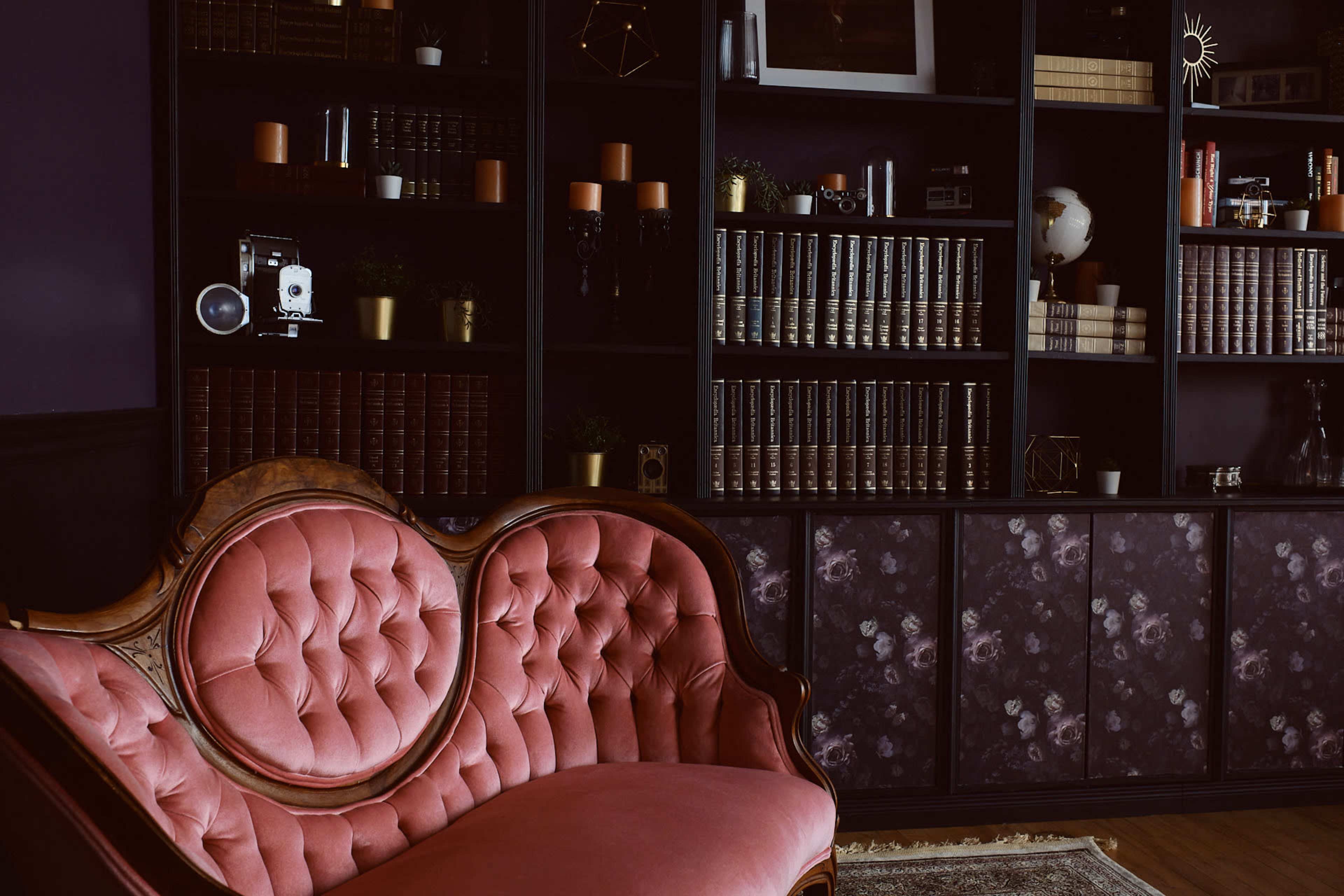 A vintage pink velvet sofa in front of a dark wooden bookshelf filled with books and decorative items.