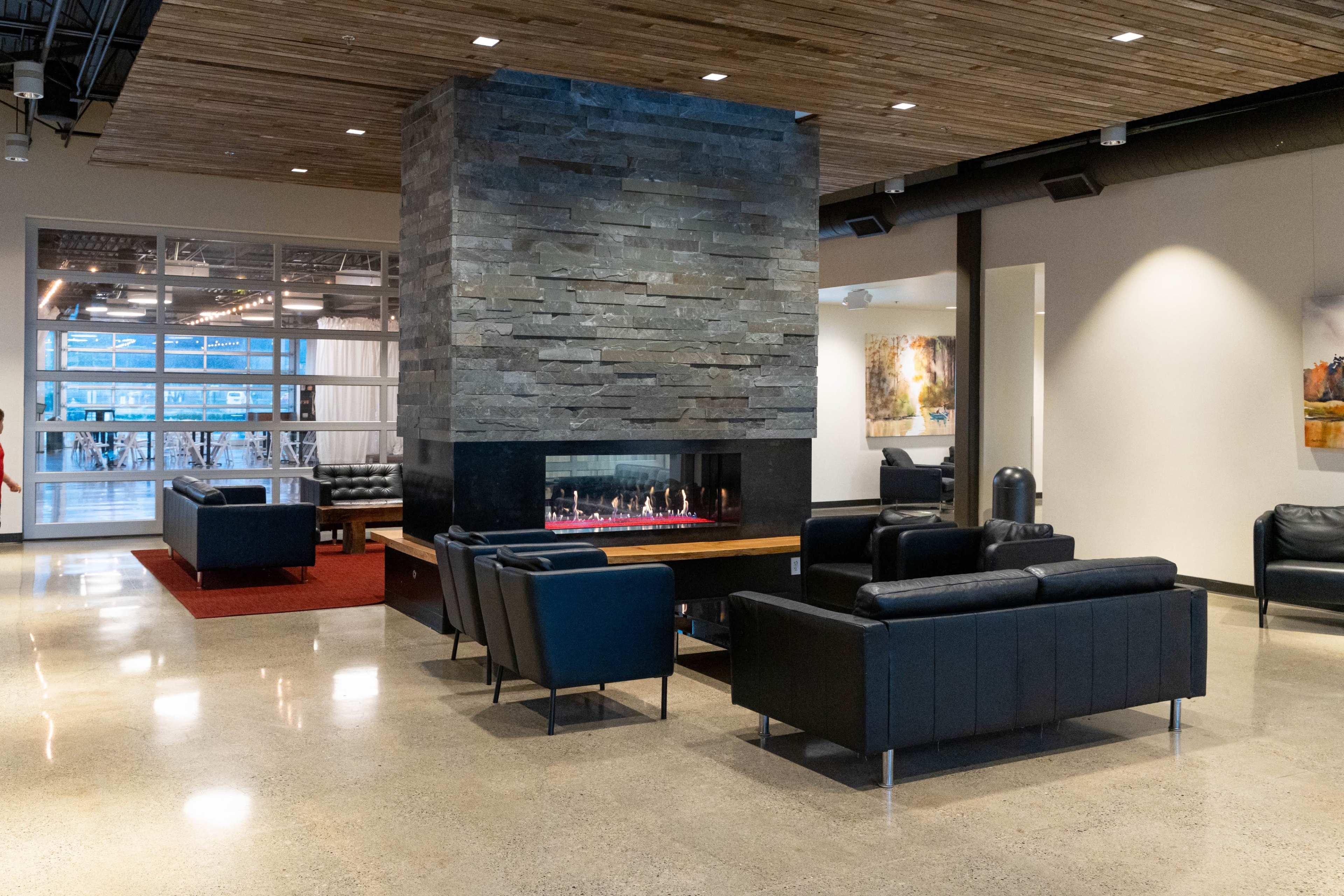 A modern lounge area with leather sofas arranged around a central stone fireplace, featuring a polished concrete floor and large windows.