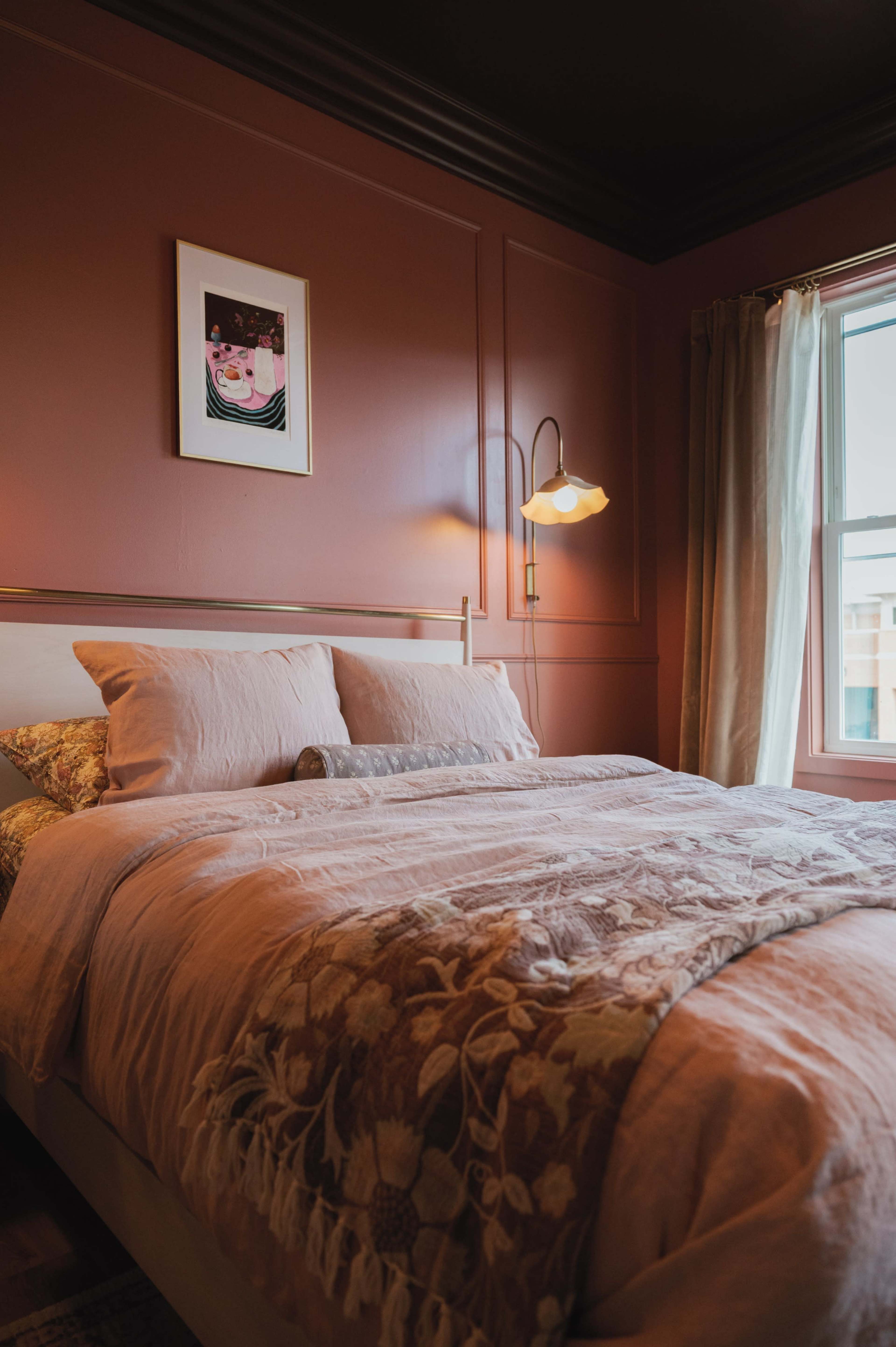 The image shows a cozy bedroom with a pink bedspread, a decorative throw blanket, and a piece of artwork on the wall beside a window.