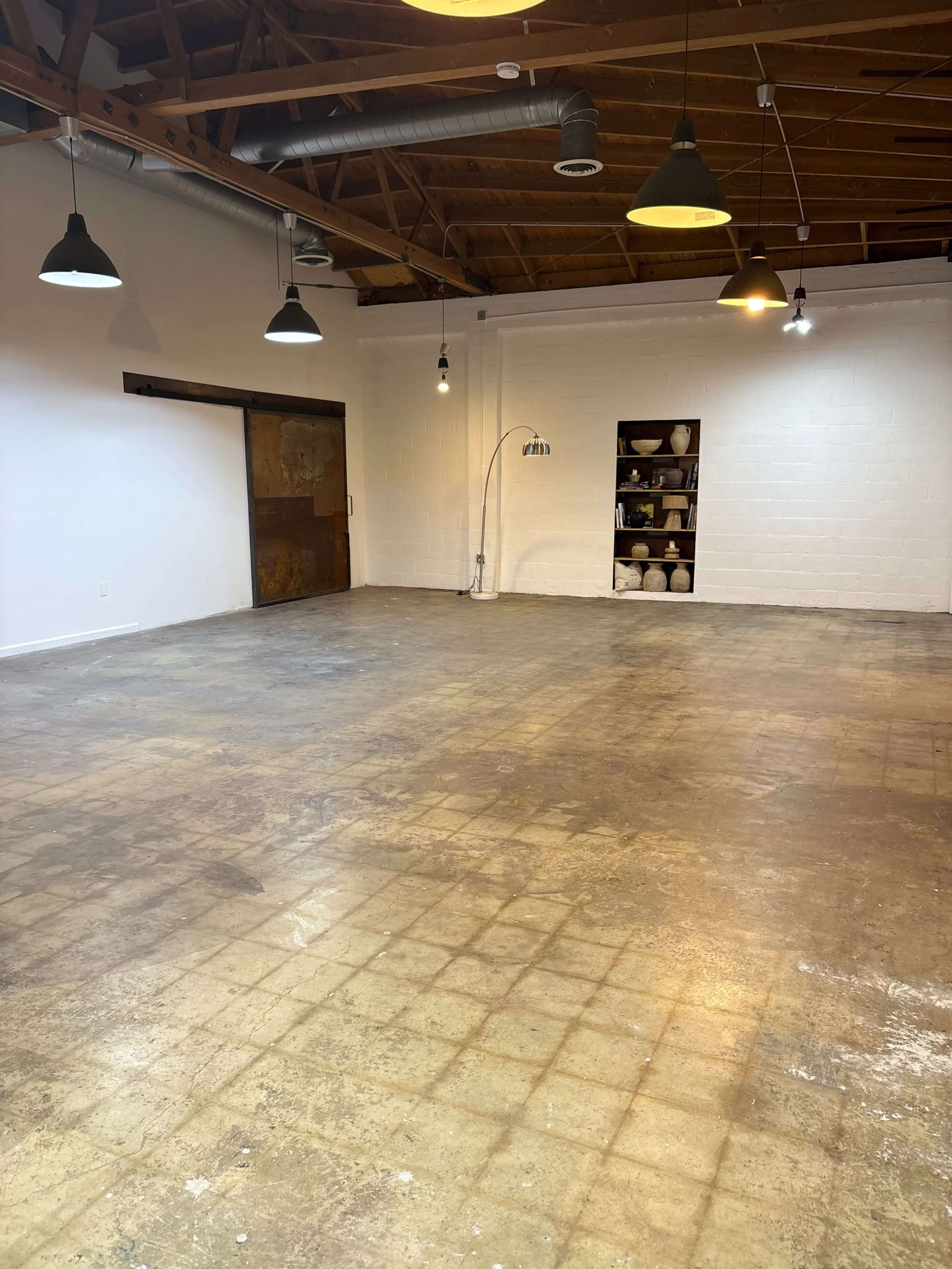 The image shows a spacious, empty room with wooden beams on the ceiling and a concrete floor, featuring a small shelf of pottery against one wall.