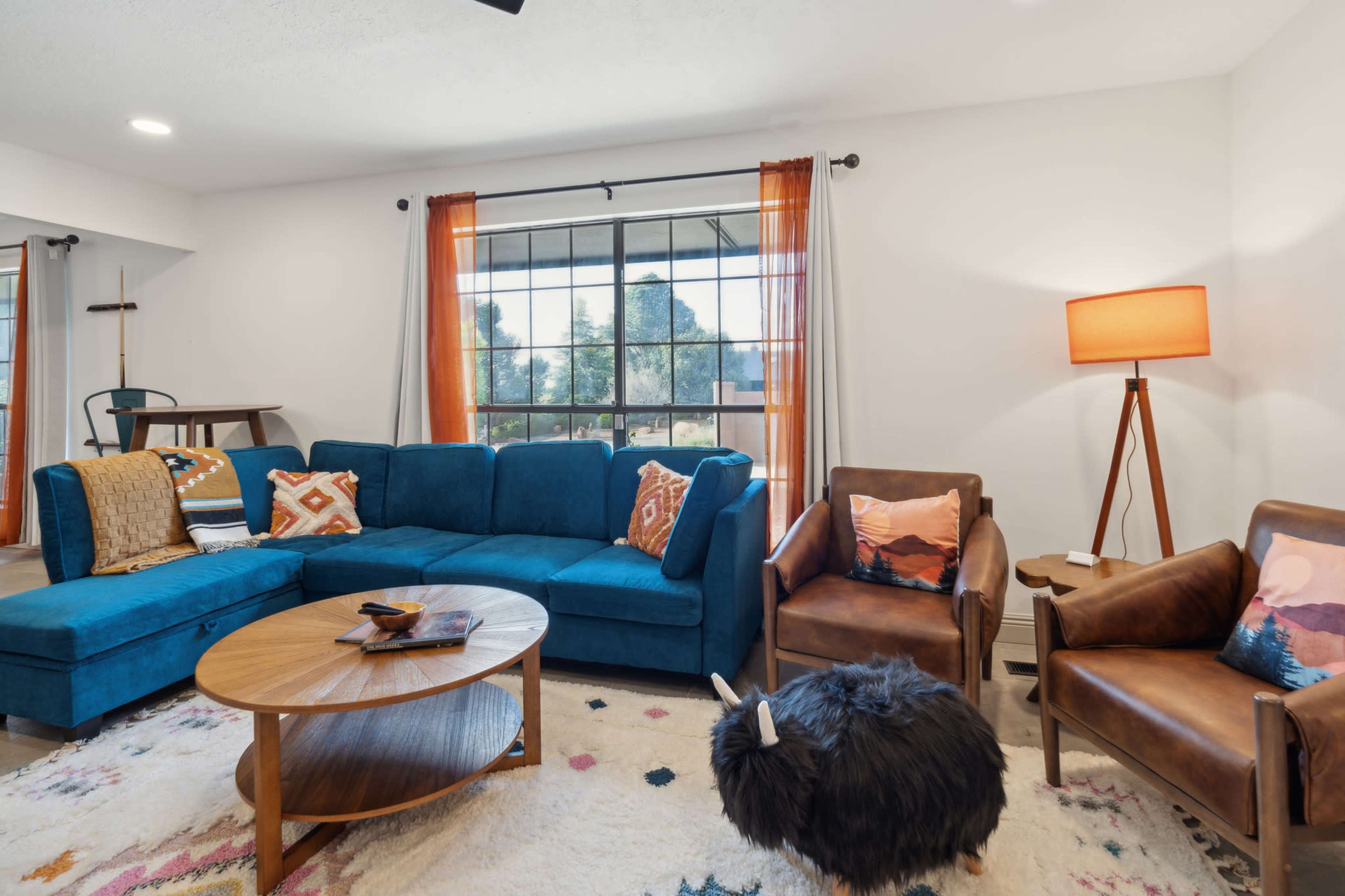 A living room with a blue sectional sofa, two brown armchairs, a wooden coffee table, and a large window dressed with orange curtains.