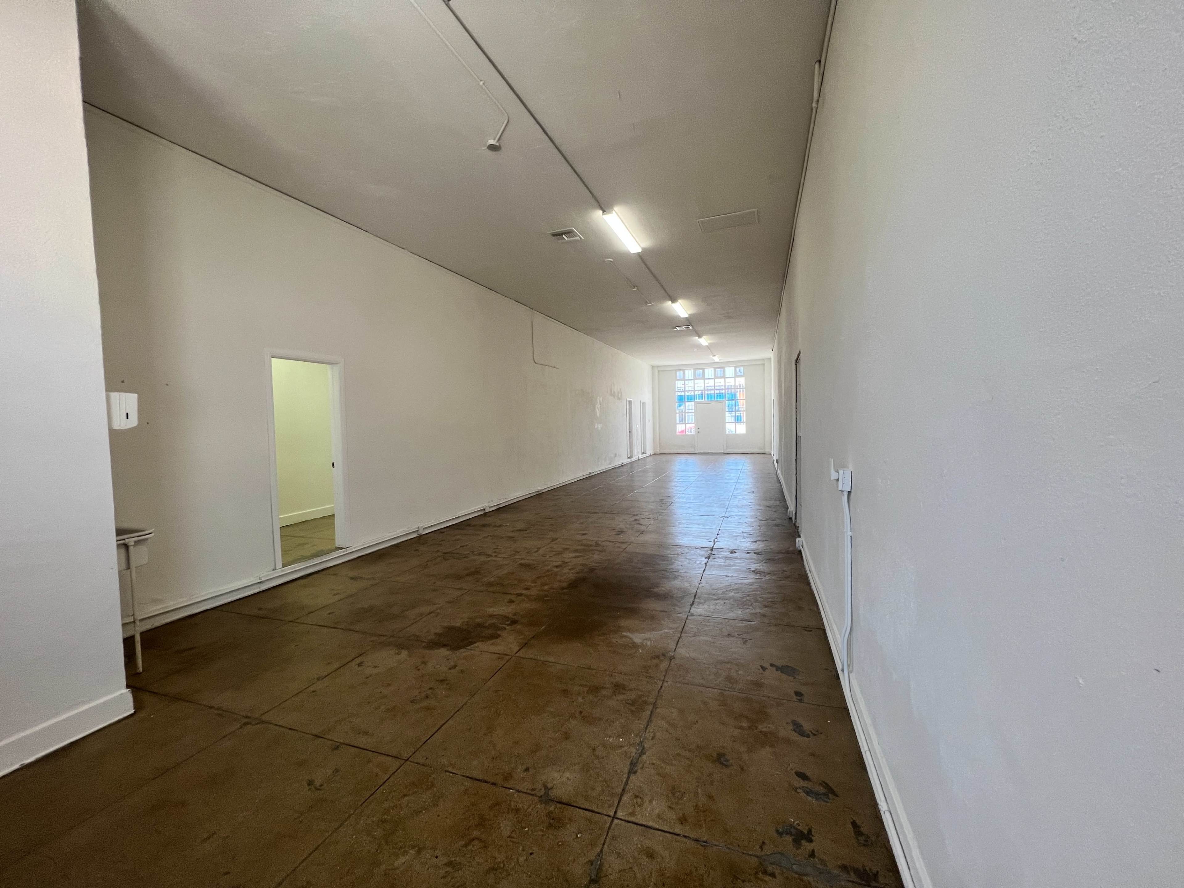 The image shows a long, empty hallway with a concrete floor and white walls, leading to a door at the far end.