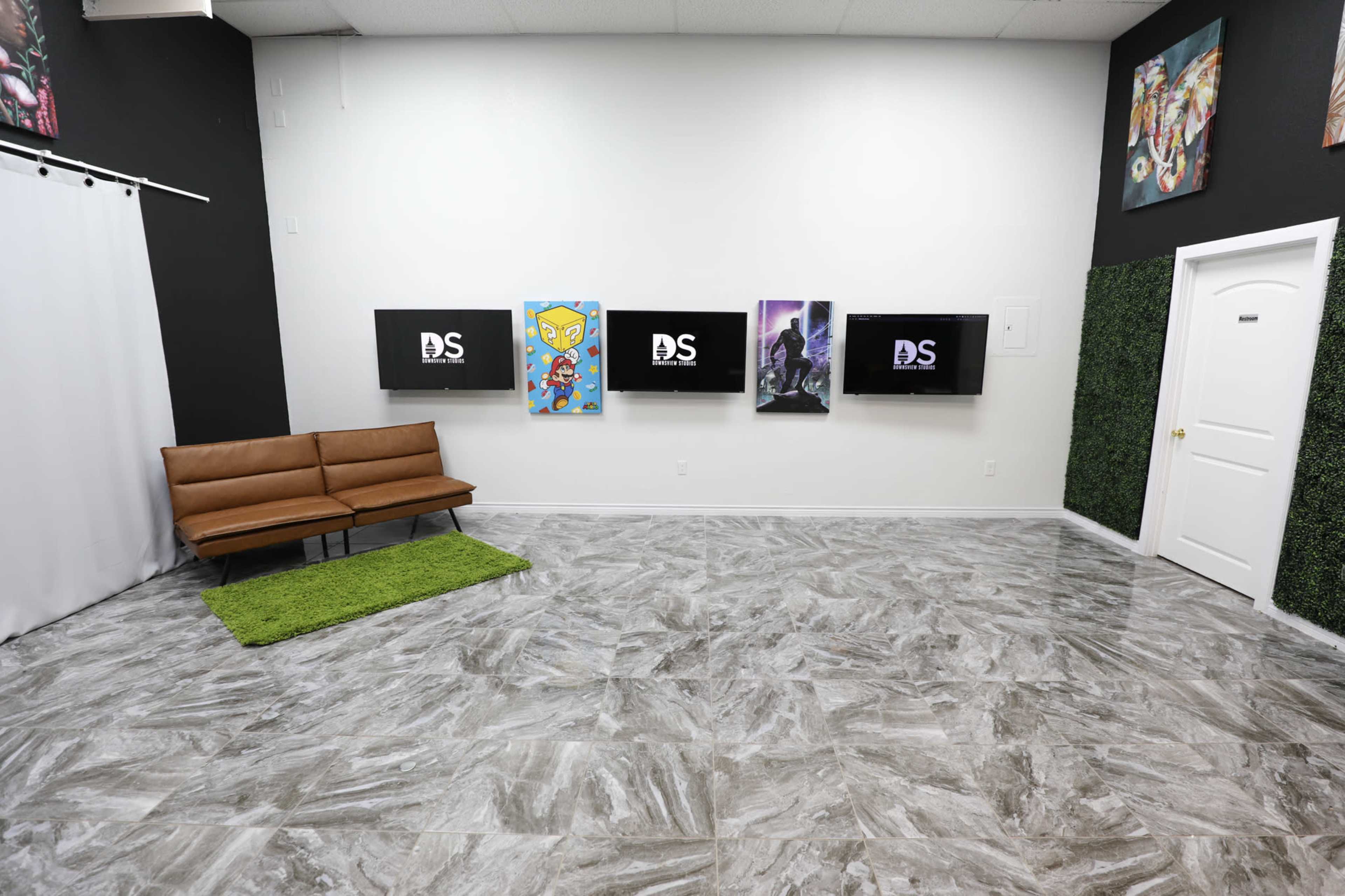 A sparsely furnished room with a brown sofa, a green rug, and three wall-mounted televisions displaying graphics, along with artwork on the walls.