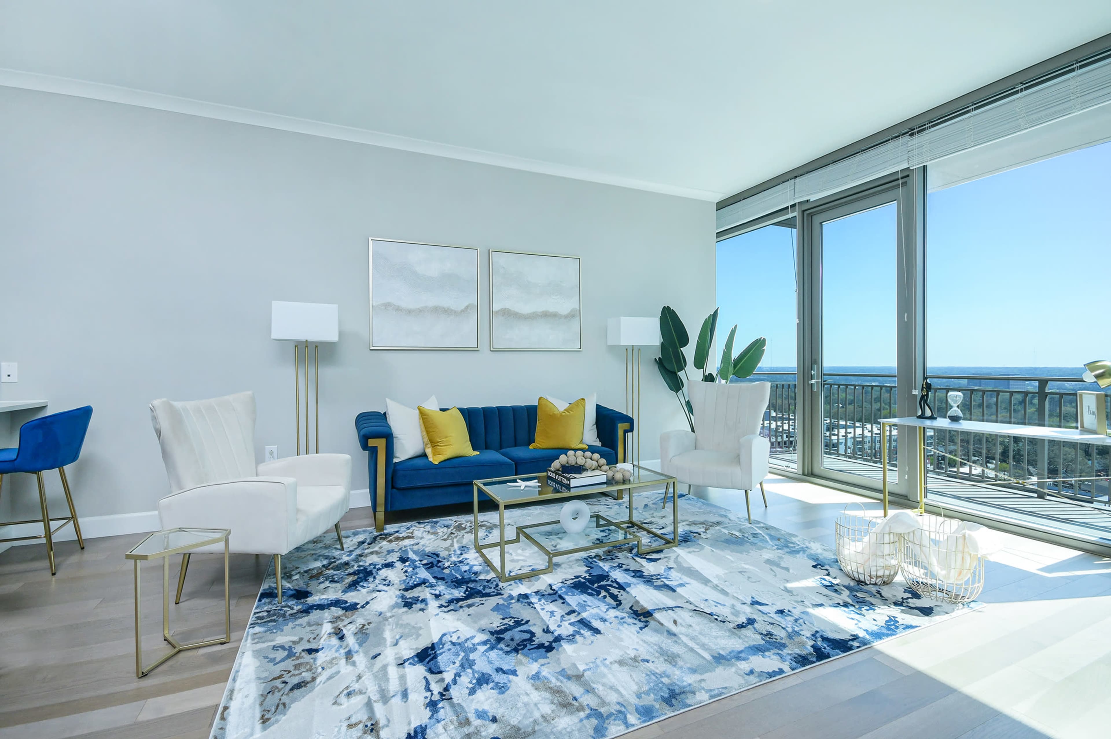 A modern living room features a blue sofa with yellow cushions, two white armchairs, a glass coffee table, and large windows with a view.