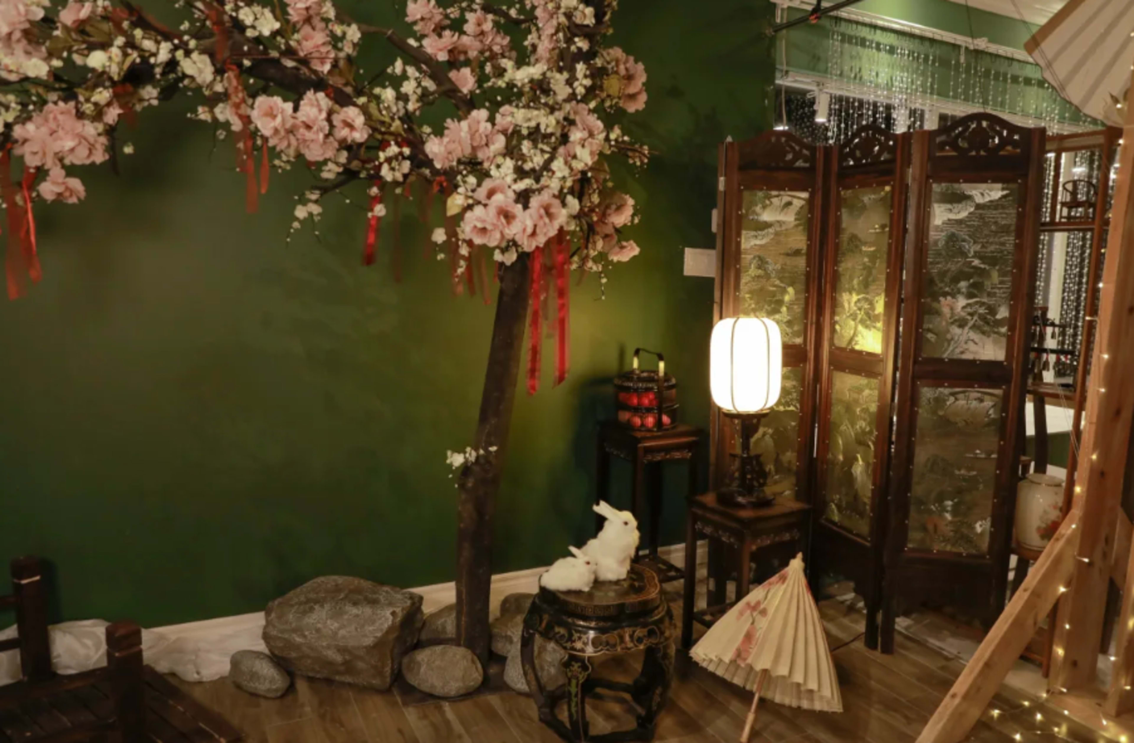 The scene features a decorative corner with a flowering tree, a traditional folding screen, a lantern, and a small table with a decorative item, all set against a dark green wall.