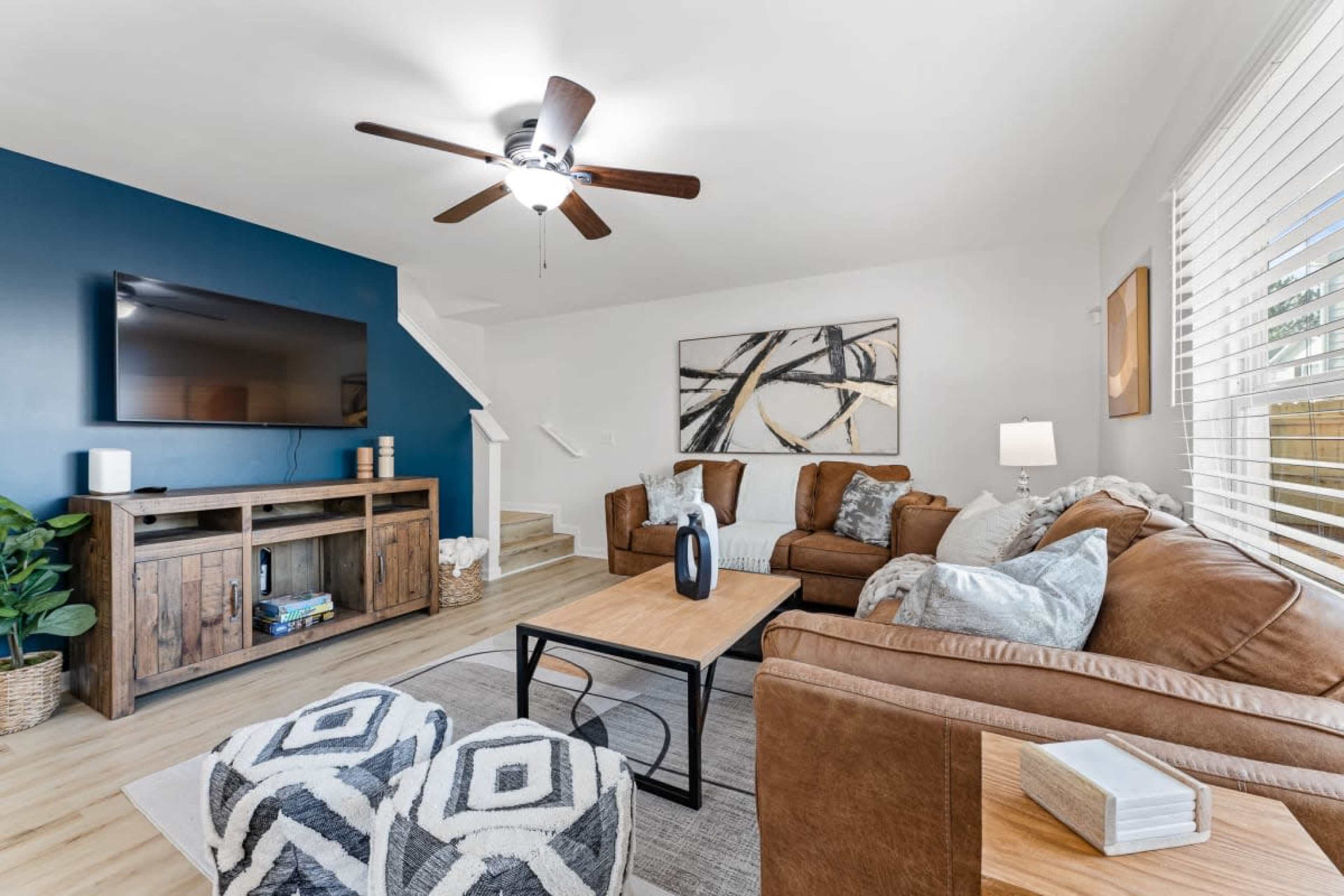 A living room features a brown suede sofa set, a wooden TV stand, and a coffee table, all arranged around a light-colored rug with patterned ottomans.