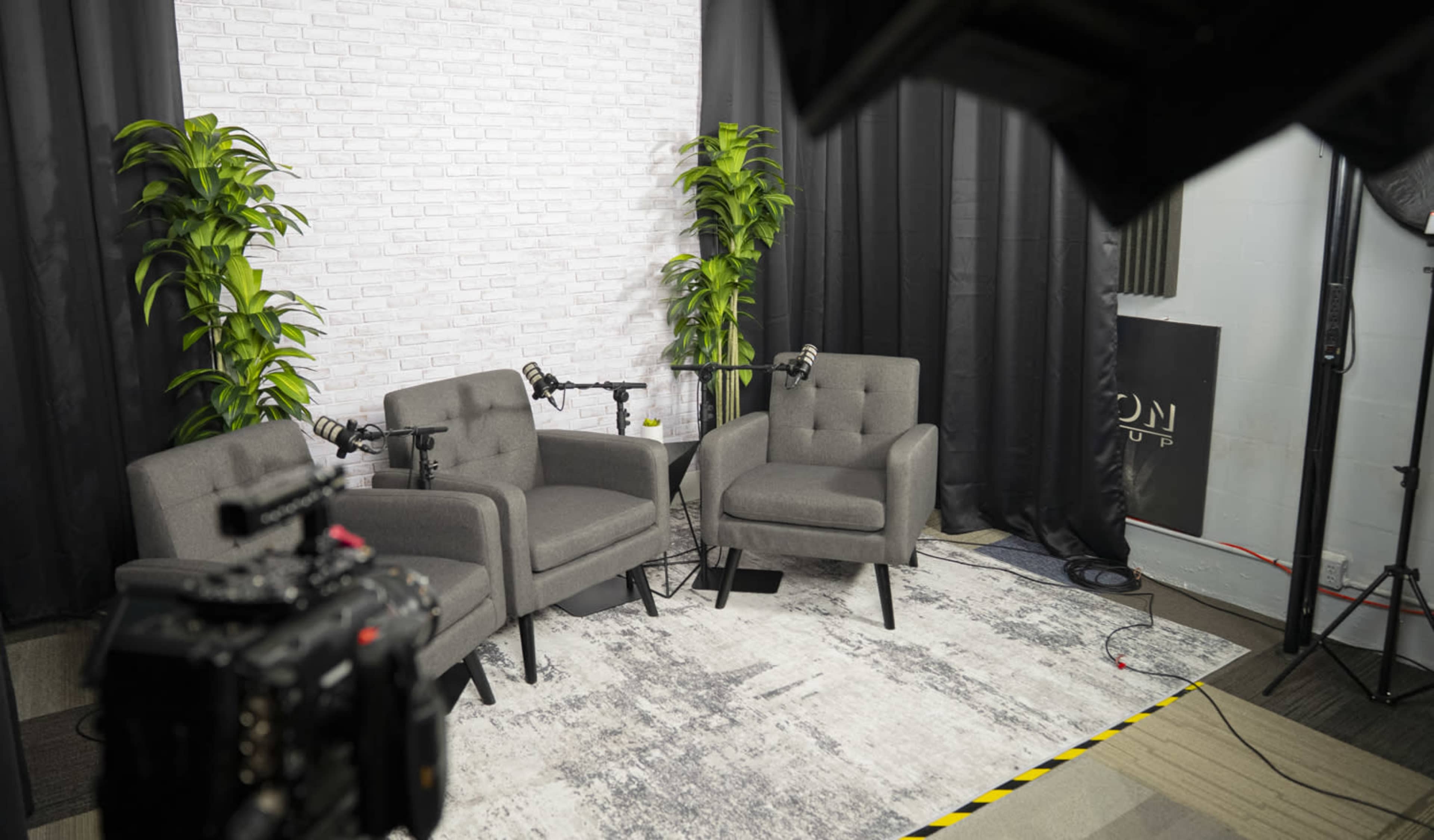 The image shows a minimalist podcast setup with two gray armchairs placed on a patterned rug, microphones positioned on each chair, and a backdrop of a white brick wall with plants on either side.