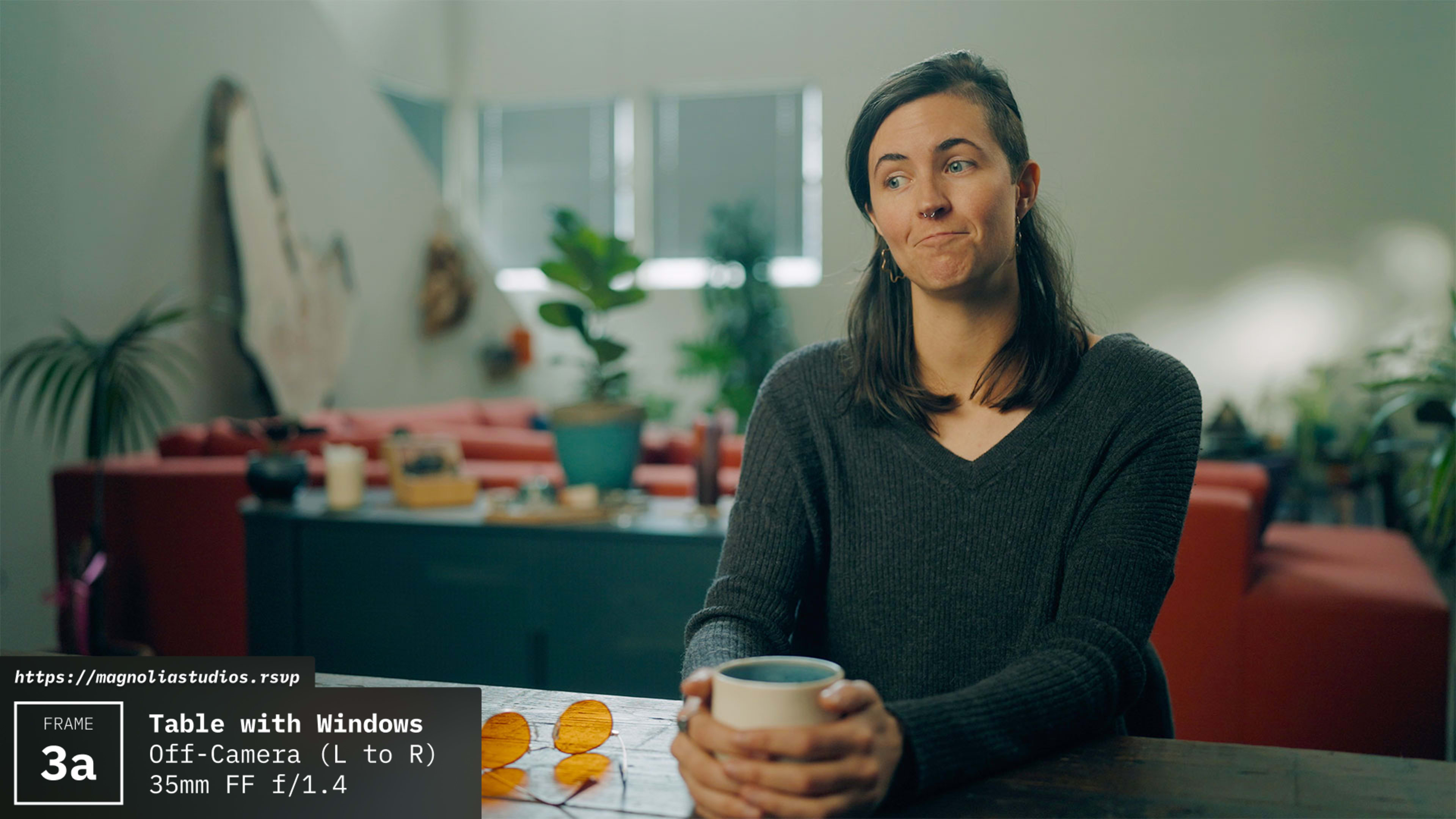 A woman with shoulder-length hair sits at a table holding a cup, with a green plant and a sofa in the background.