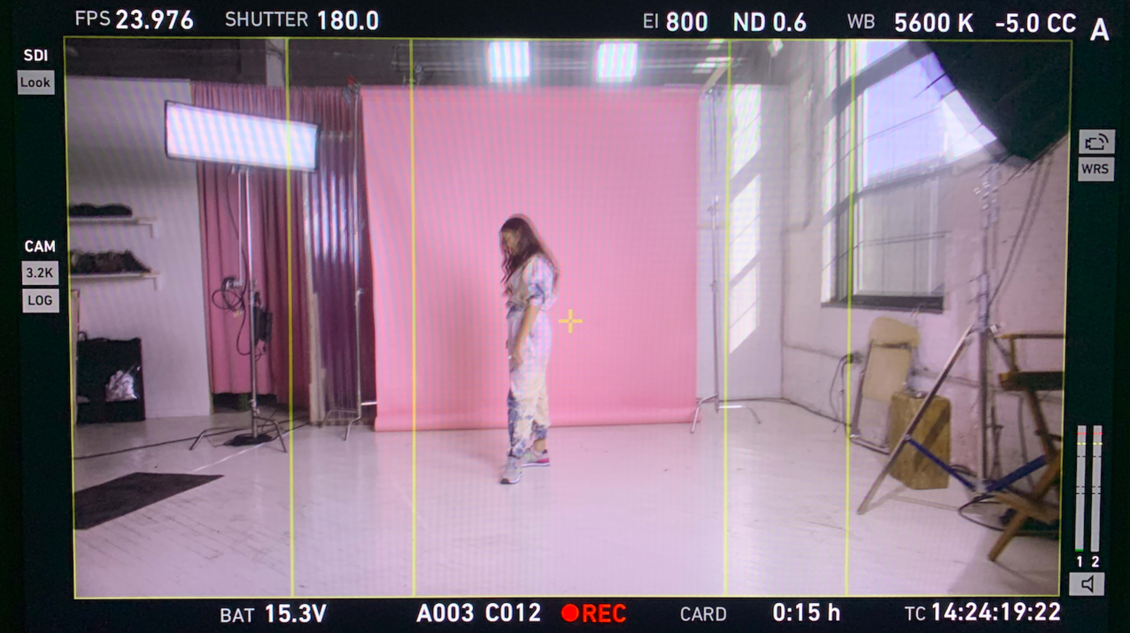 A model stands in a photography studio with a pink backdrop, bright lights, and a computer monitor displaying camera settings.