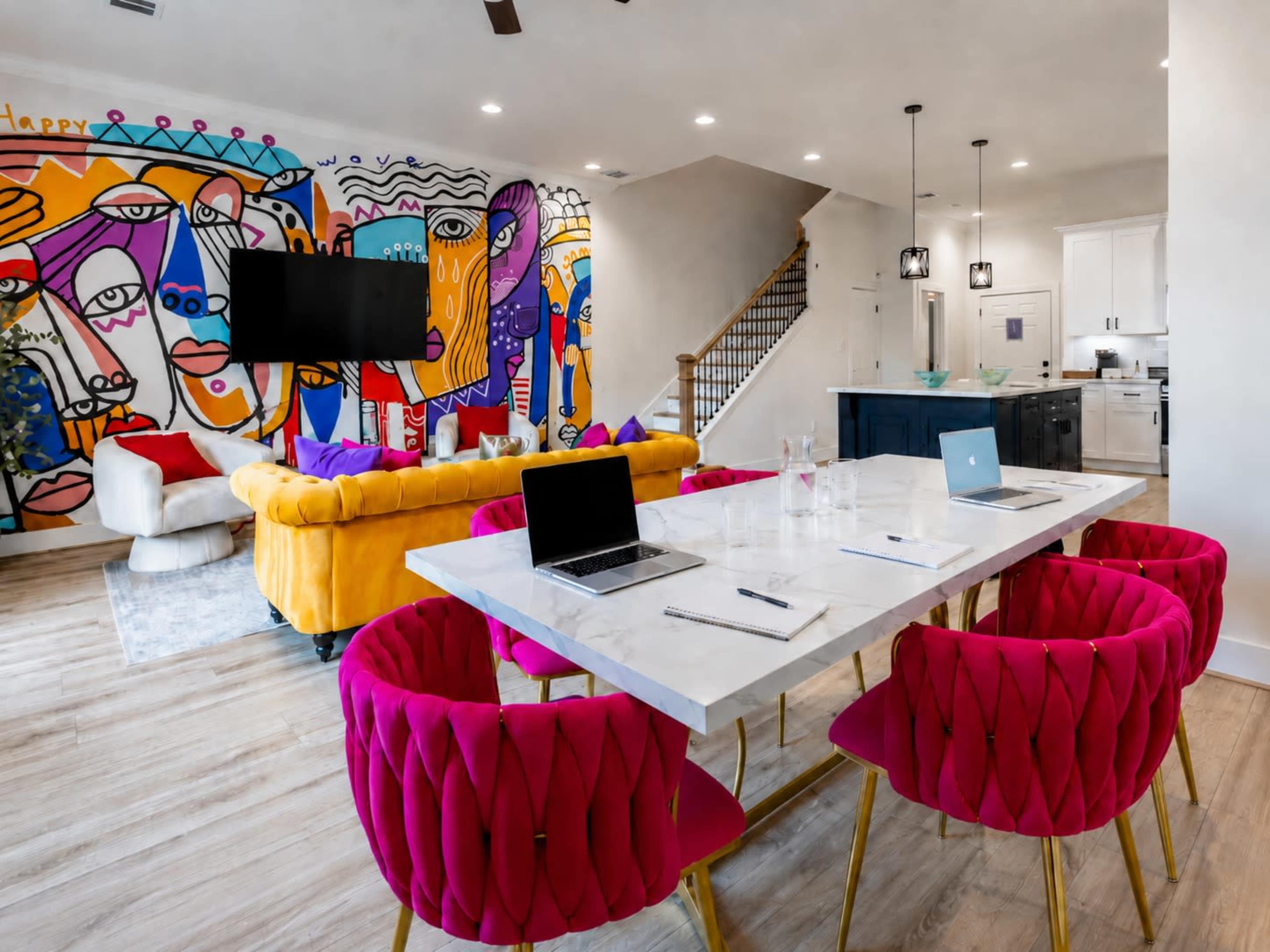 The image shows a modern living space with a colorful mural on the wall, a yellow sofa, pink chairs around a marble table, and a kitchen in the background.