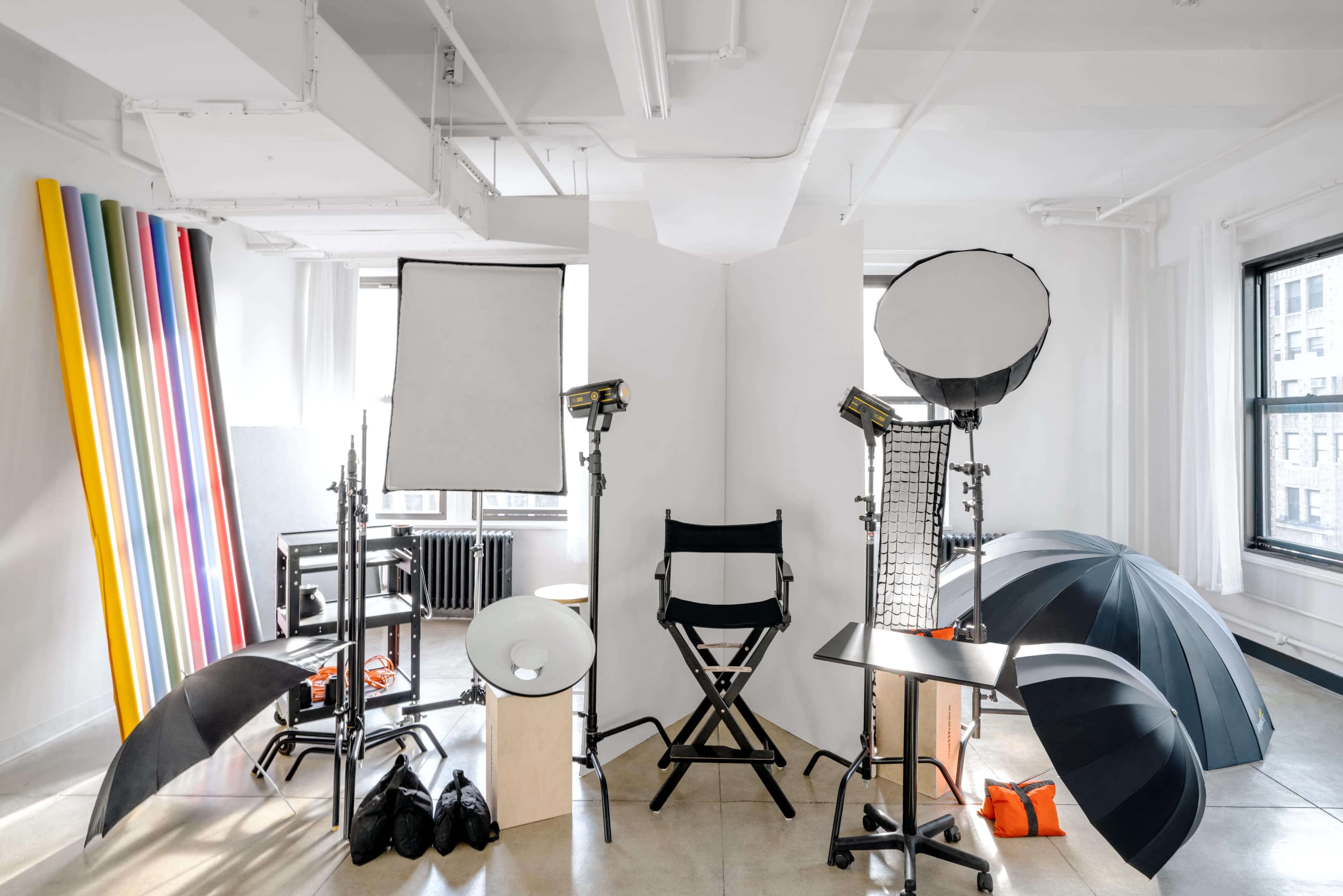 A photography studio setup features multiple light sources, reflectors, and a director's chair surrounded by various studio equipment.