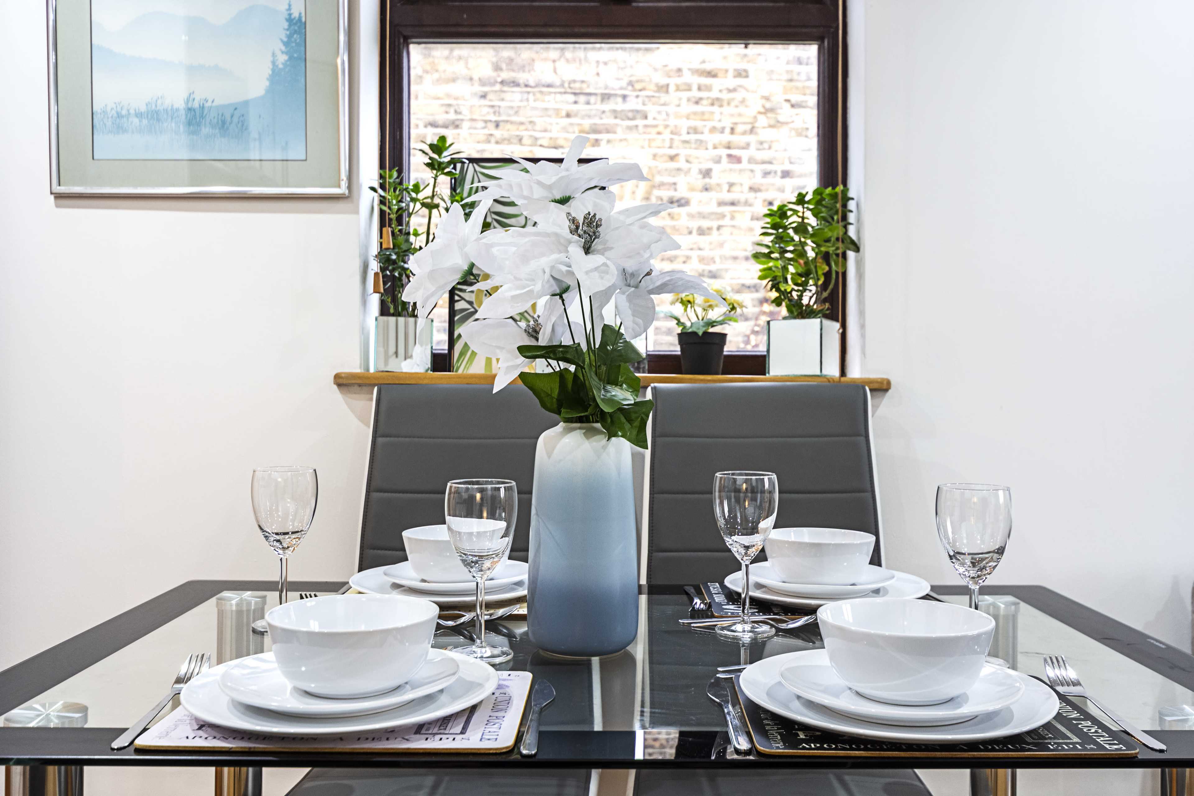 A dining table is set with white dishes and glassware, featuring a vase of white flowers and greenery in the background.
