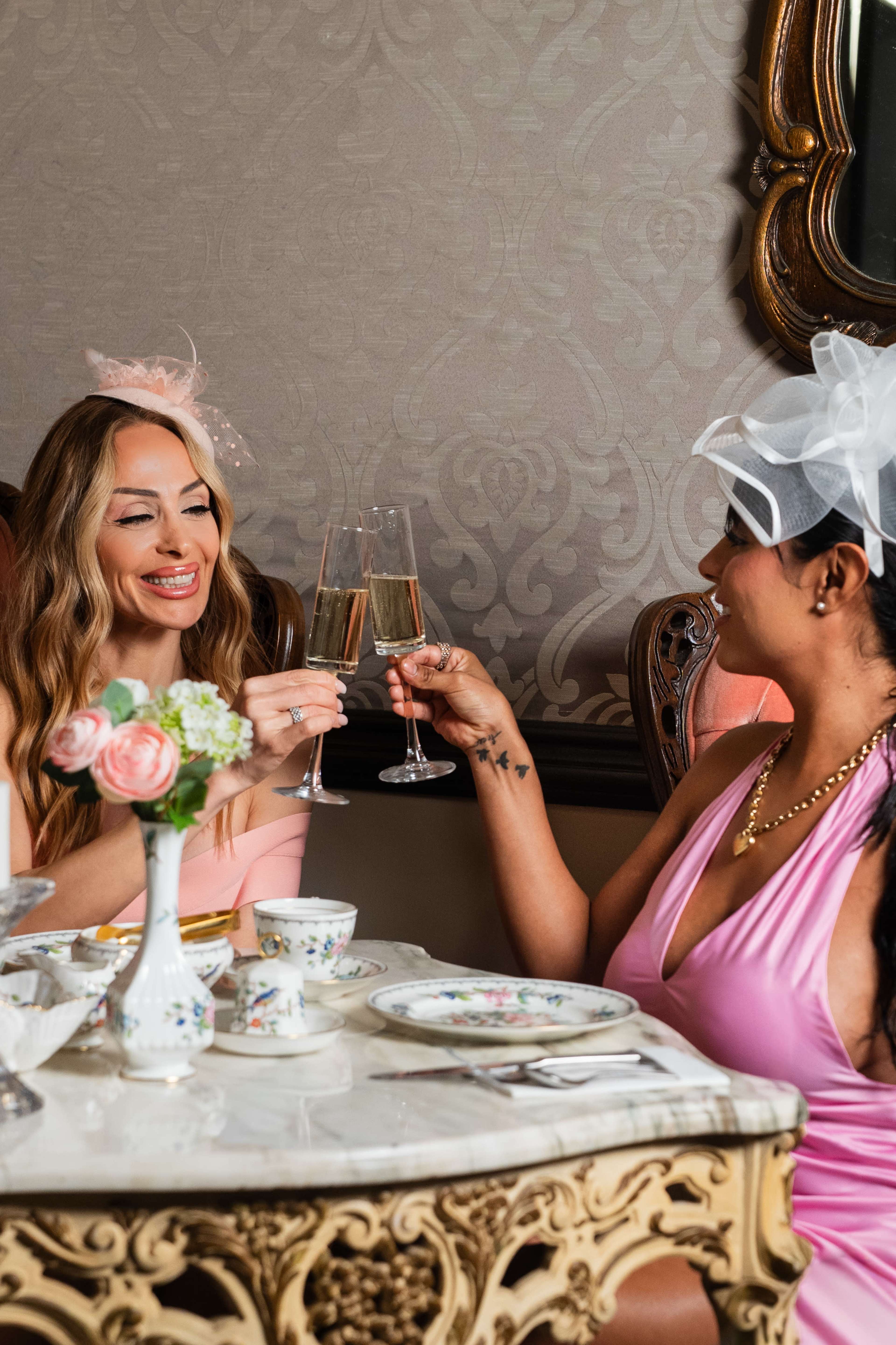 Two women, one in a pink dress and another in a light pink top, are toasting with champagne glasses at an elegantly set table adorned with floral arrangements and fine china.