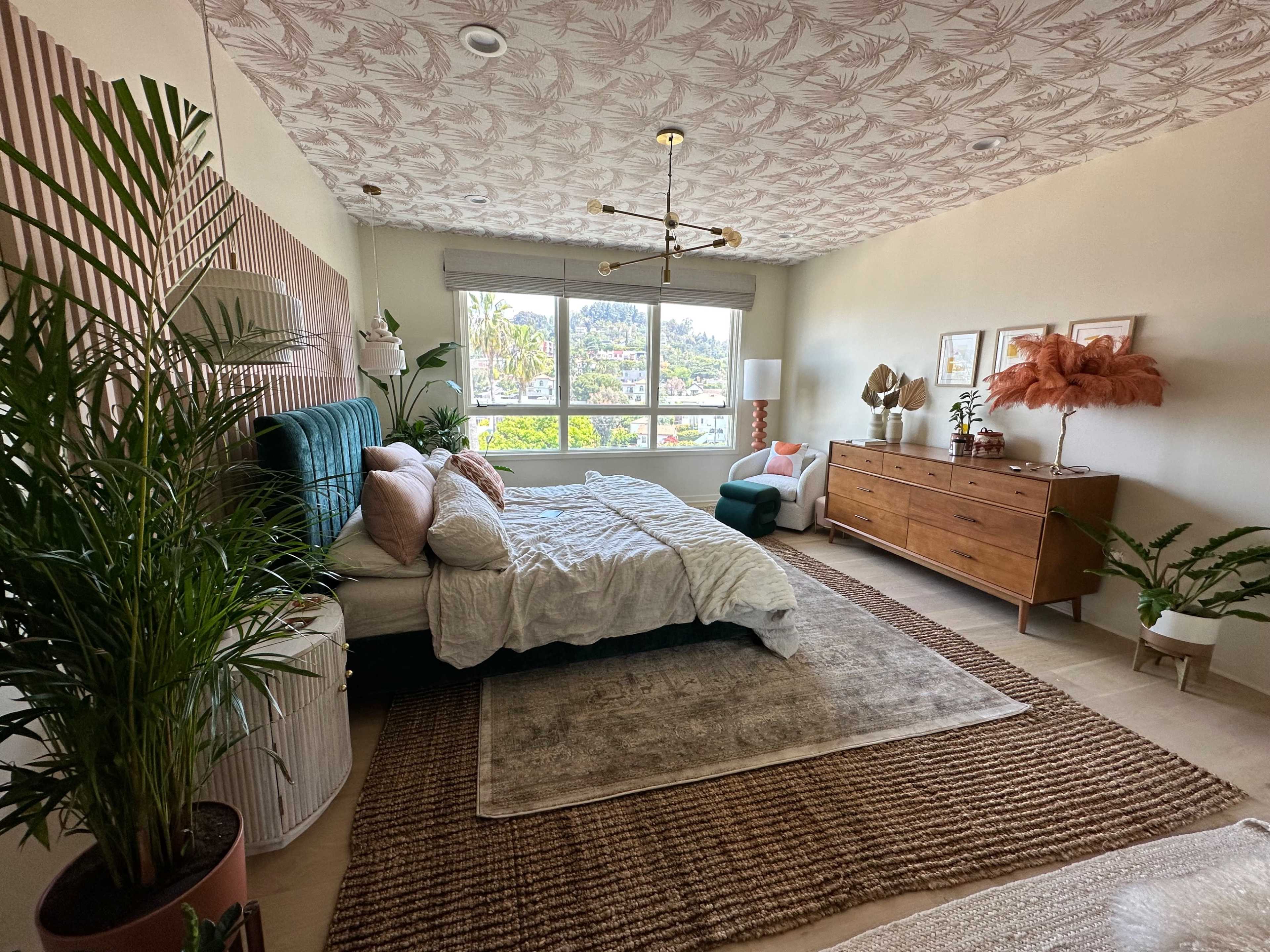 A spacious bedroom features a large bed with a patterned bedspread, a mid-century modern dresser, and large windows overlooking greenery.