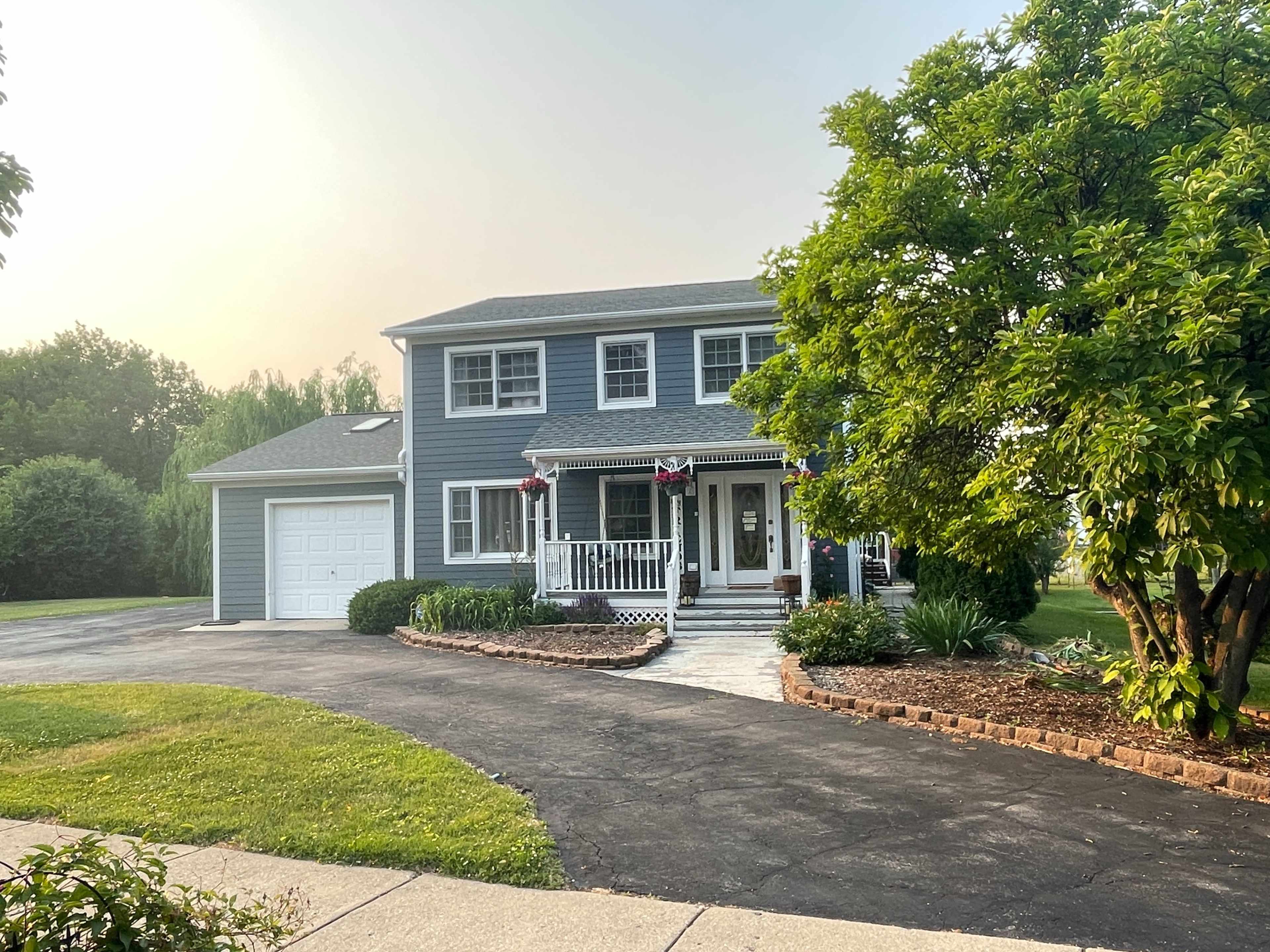 A two-story blue house with a front porch and a driveway curves around a landscaped yard.