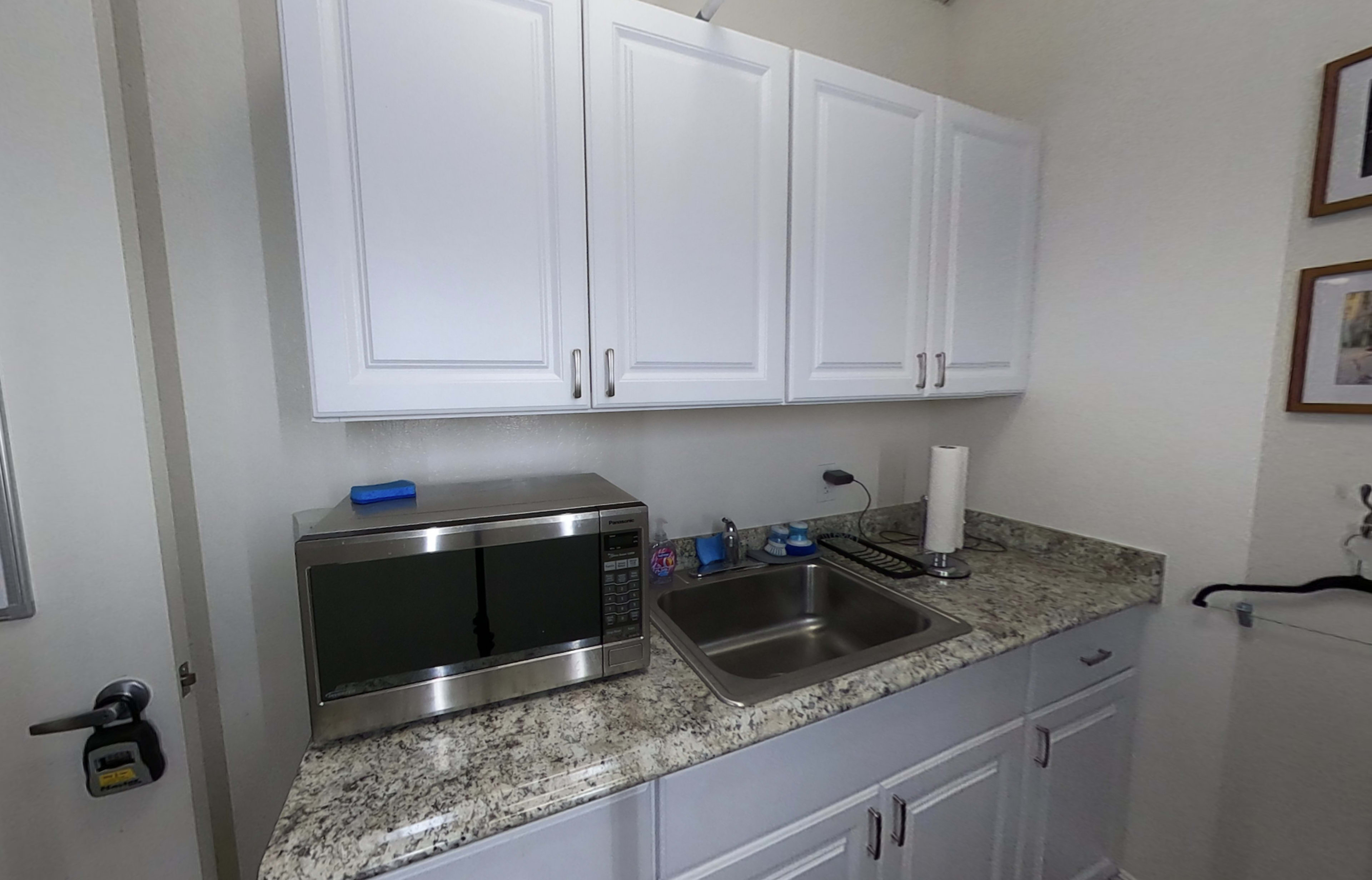 A compact kitchen area features white cabinets, a stainless steel microwave, a sink, and a countertop with various kitchen items.