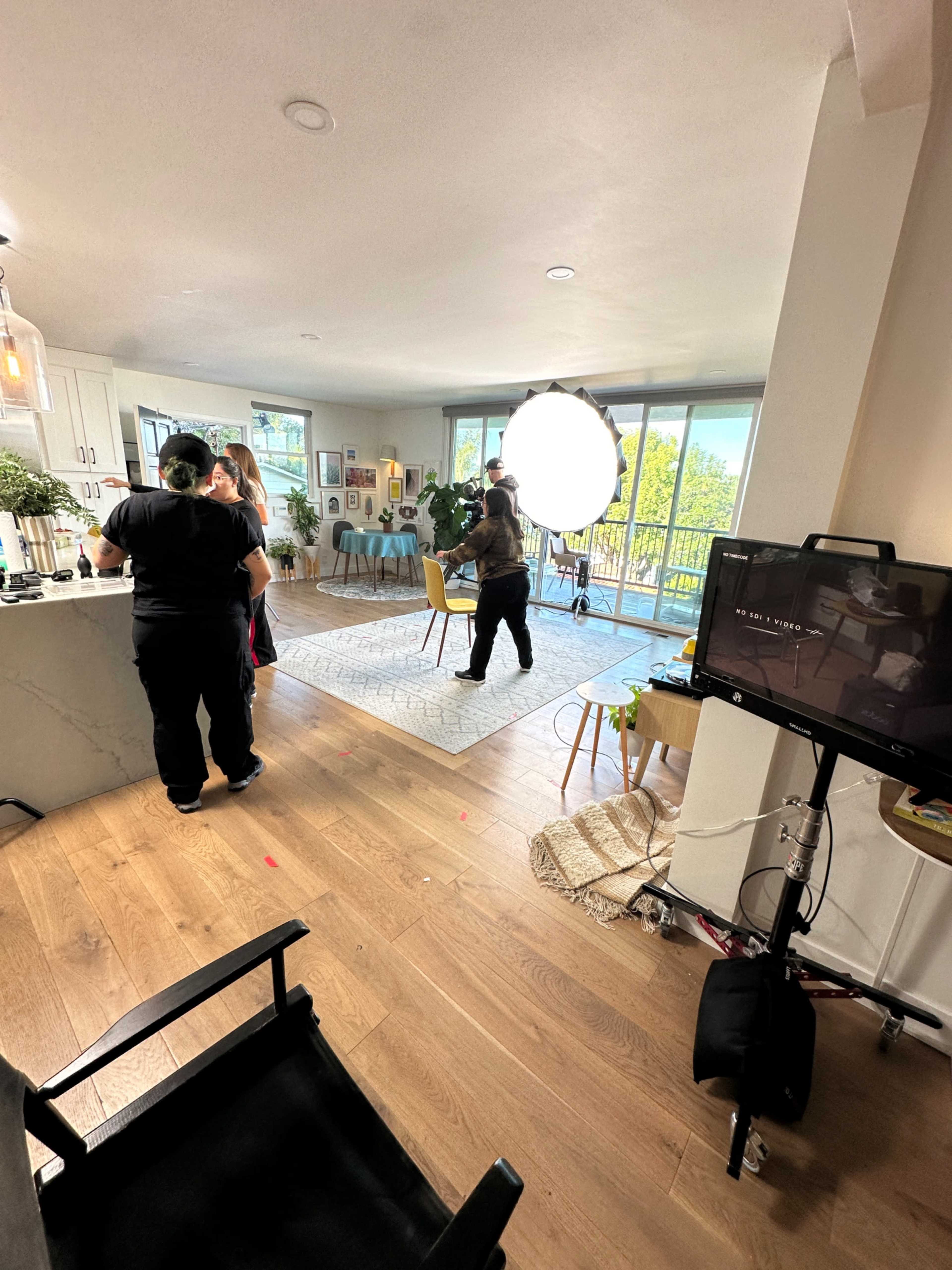 A group of people is filming in a well-lit living room, with a large softbox lighting setup and a monitor nearby.