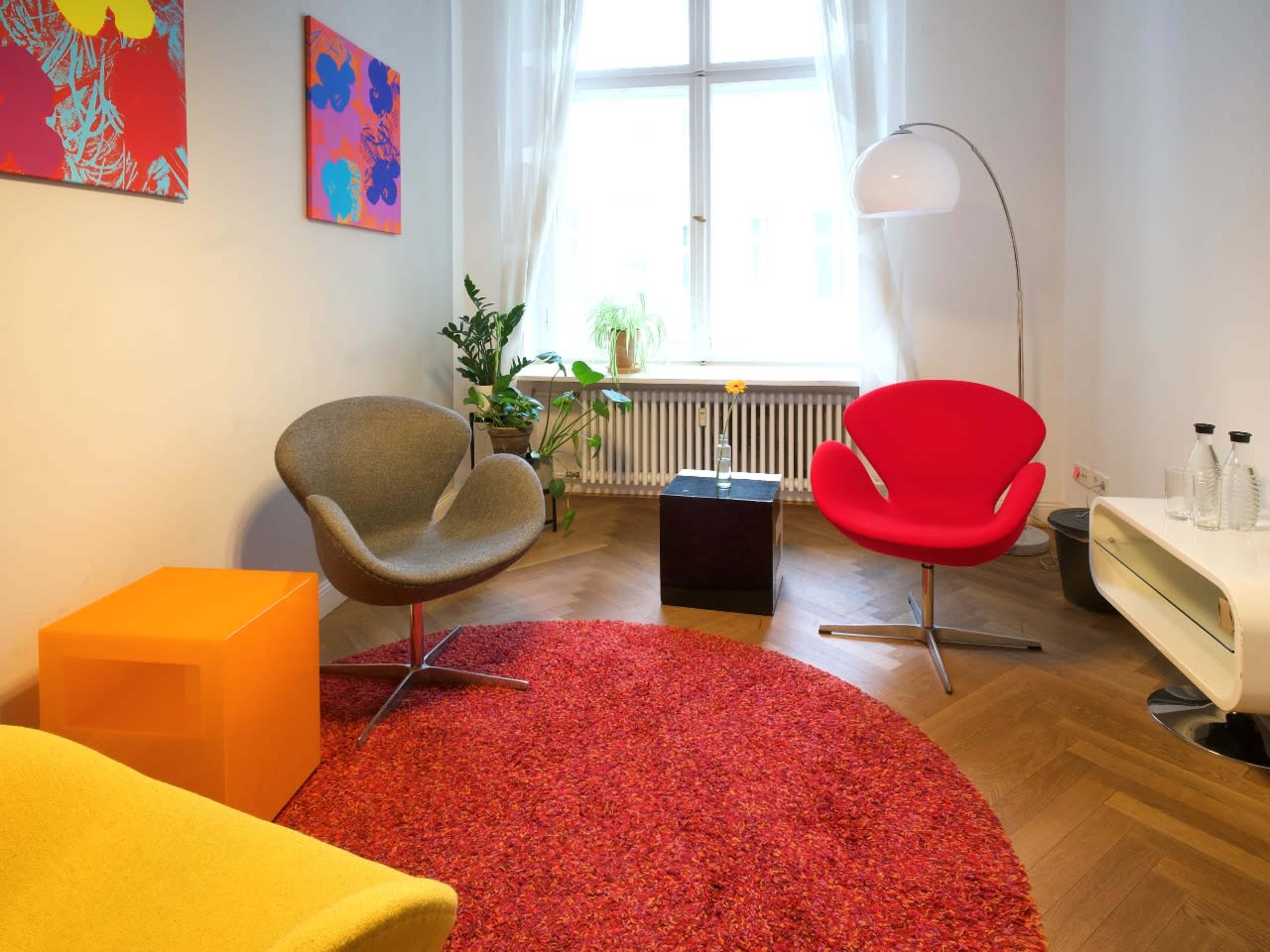 A modern, brightly colored living space featuring a red chair, a gray chair, an orange cube, and a round red rug, with a large window allowing natural light to enter.