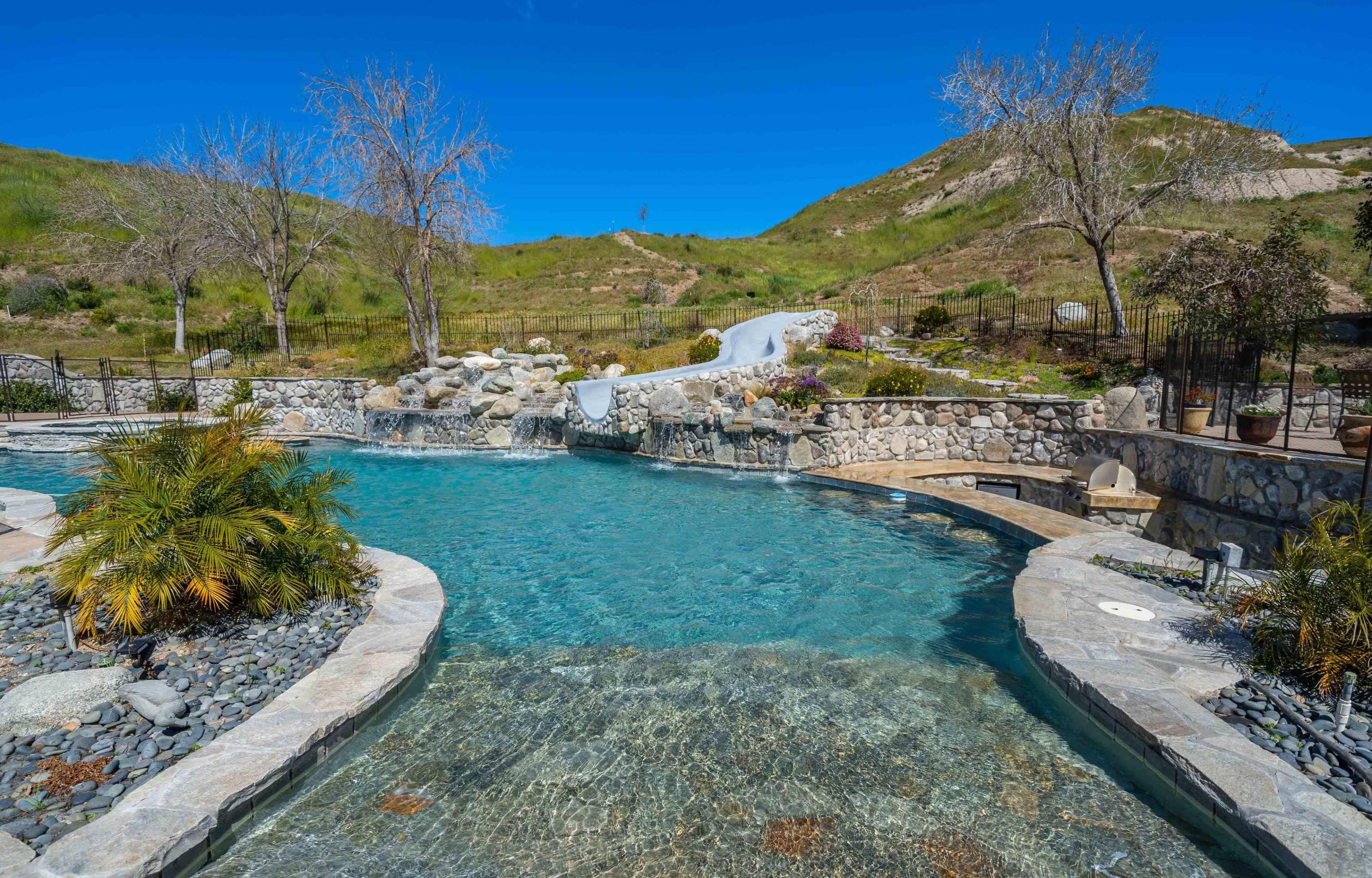 A clear, blue swimming pool is surrounded by stone pathways, landscaping, and dry trees, set against a green hillside under a bright blue sky.