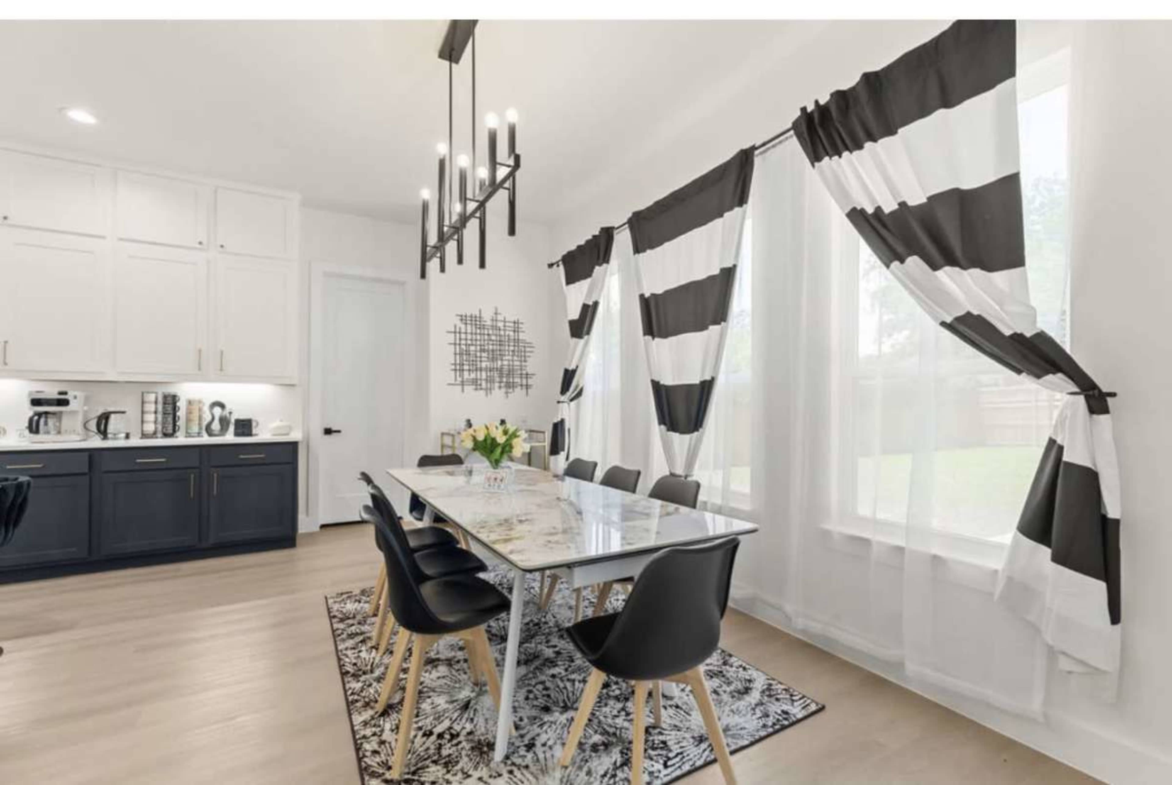 The image shows a modern dining area featuring a long table with black chairs, set against a backdrop of striped curtains and a light-colored wall.