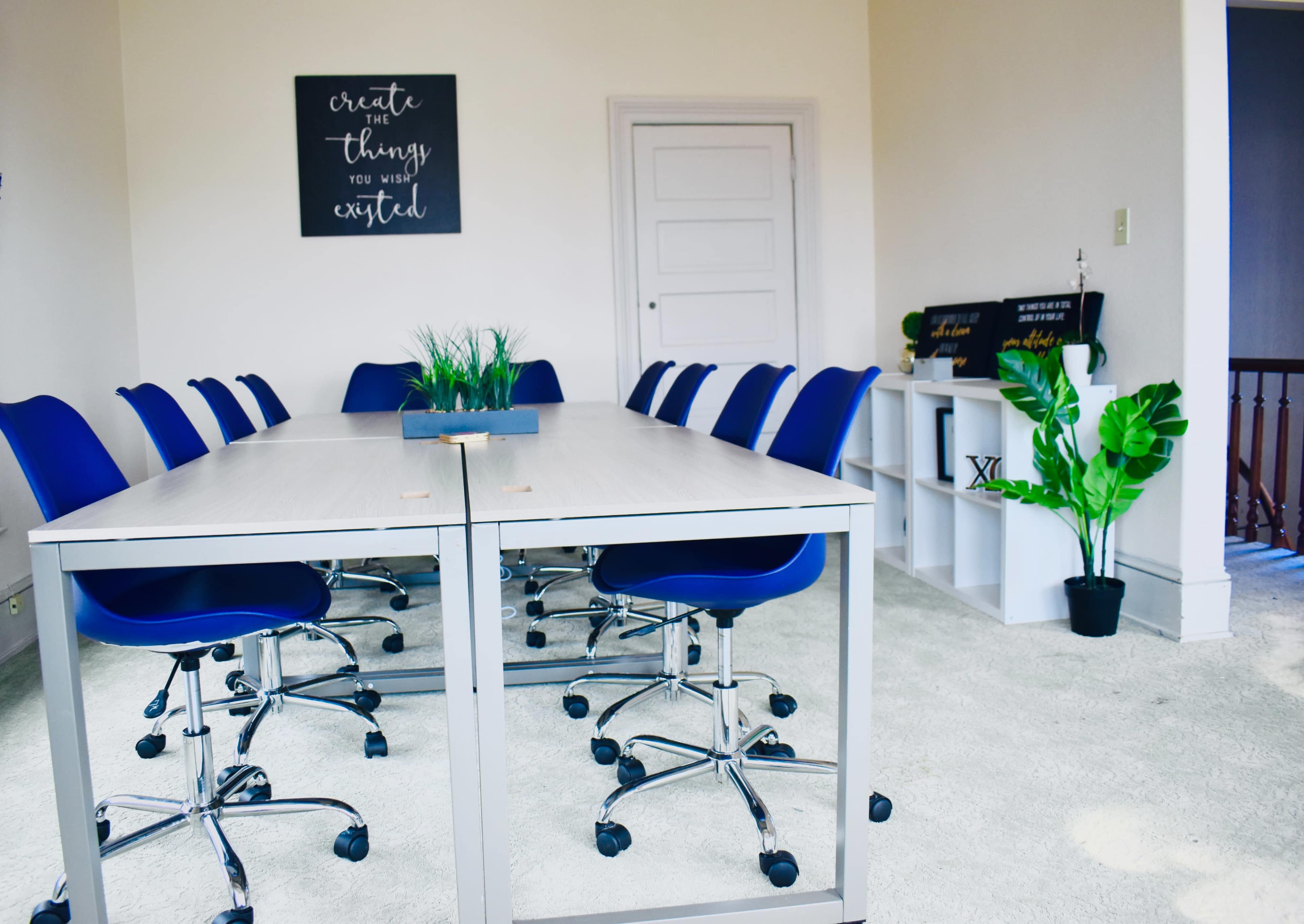A conference room features a long table surrounded by blue chairs, with greenery and motivational decor on the walls.