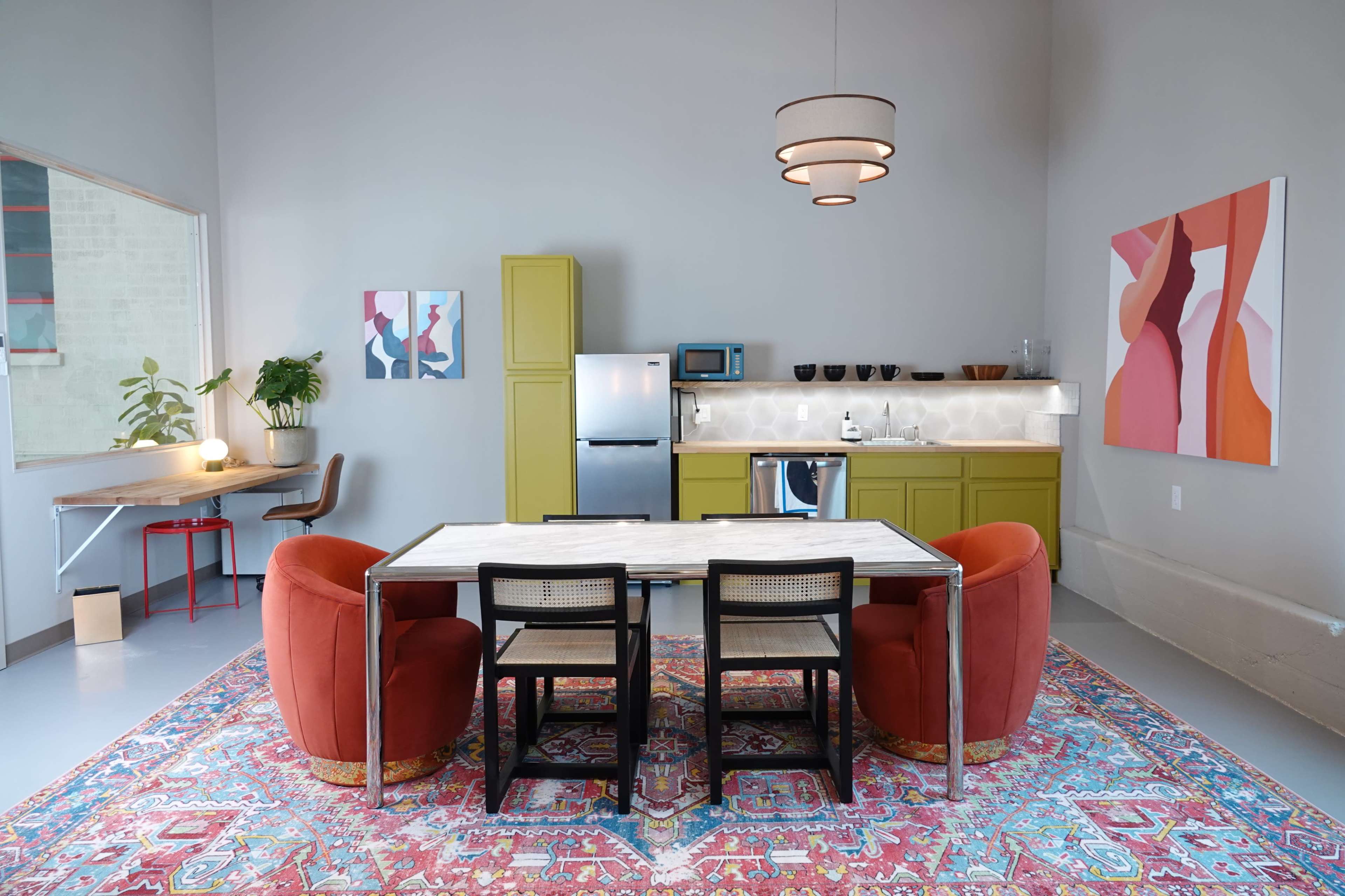 A modern kitchen and dining area features a table surrounded by black and red chairs, with a patterned rug beneath and colorful artworks on the walls.