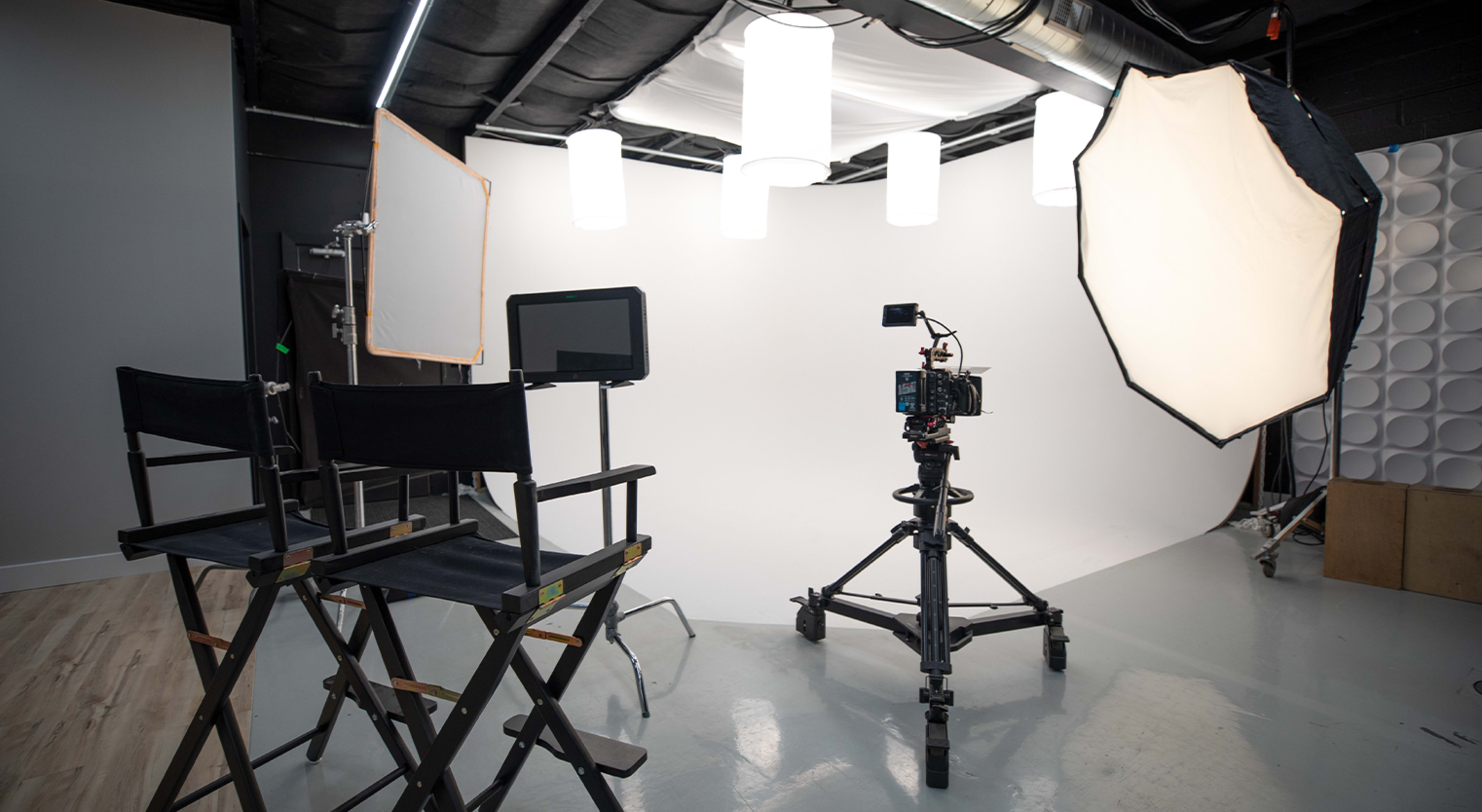 A photography studio equipped with two director's chairs, a camera on a tripod, and lighting equipment against a white backdrop.