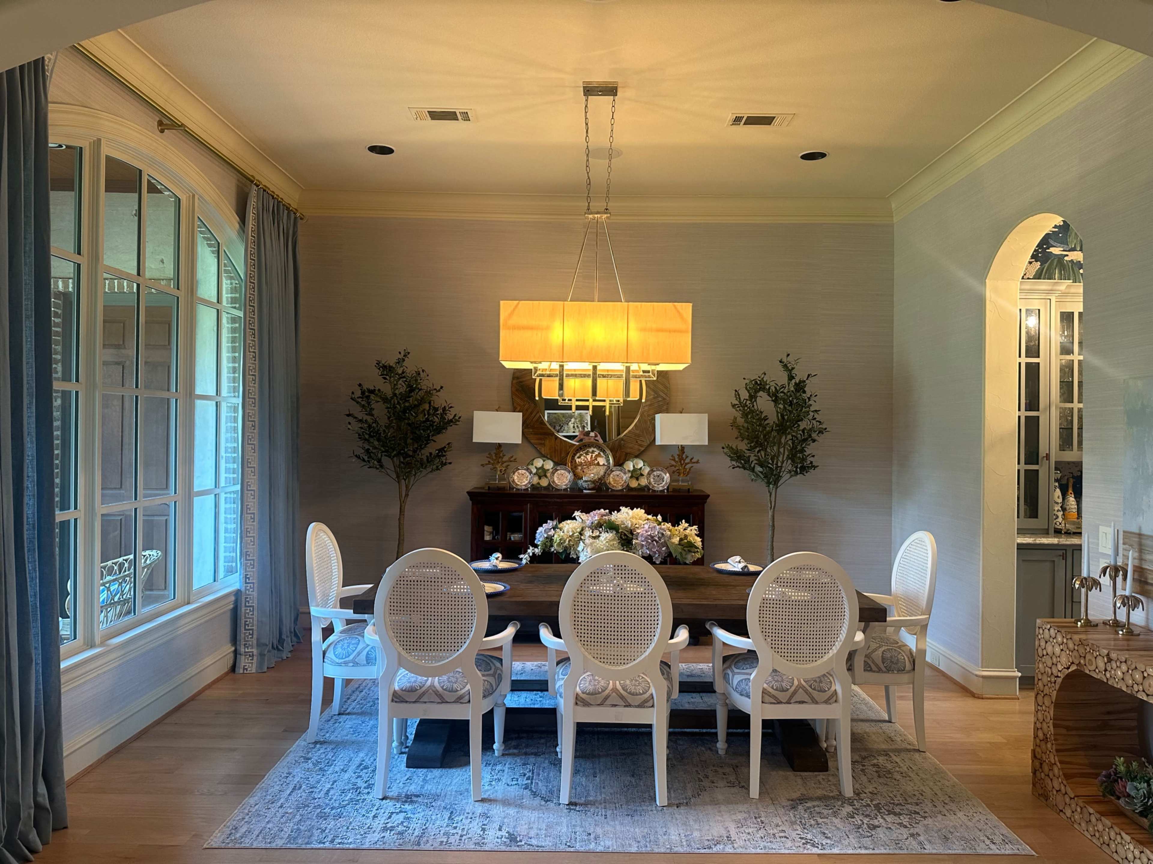 A dining room features a long table with white chairs, flanked by two potted plants, and is illuminated by a large pendant light above.