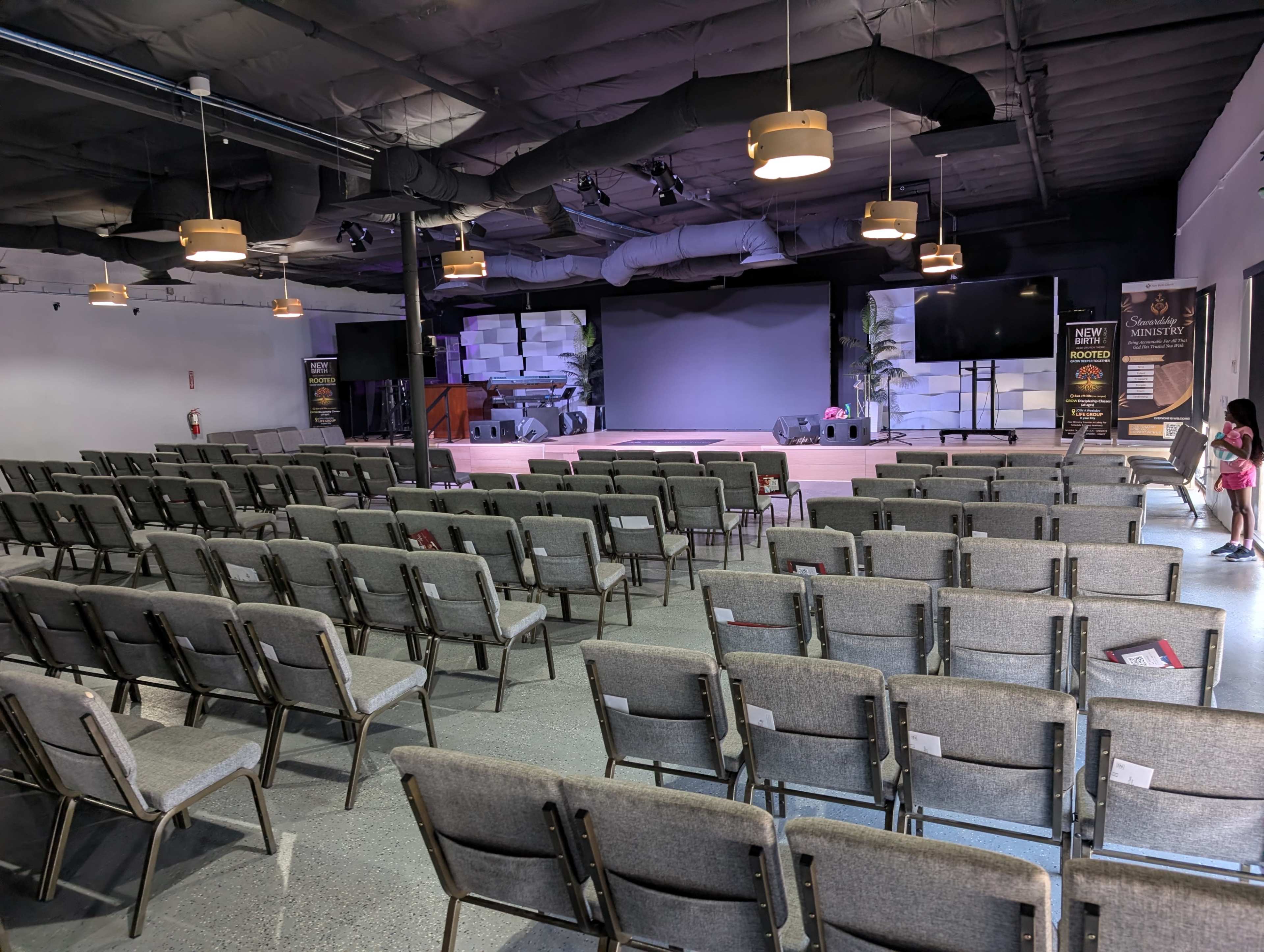 The image shows an empty auditorium with rows of gray chairs facing a stage that is illuminated by overhead lights.