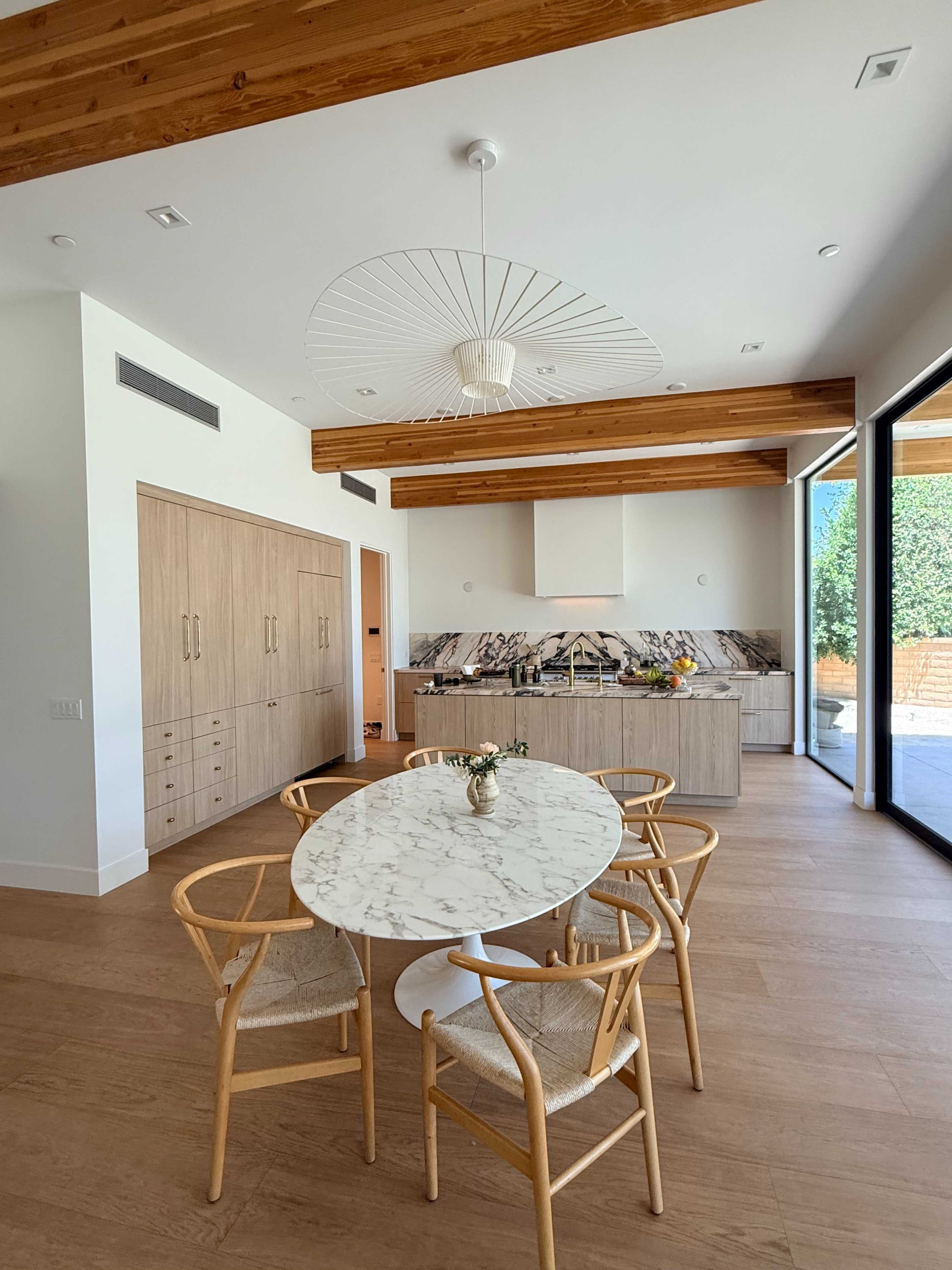 The image shows a modern kitchen and dining area with a marble table surrounded by four wooden chairs, and a large window allowing natural light to fill the space.