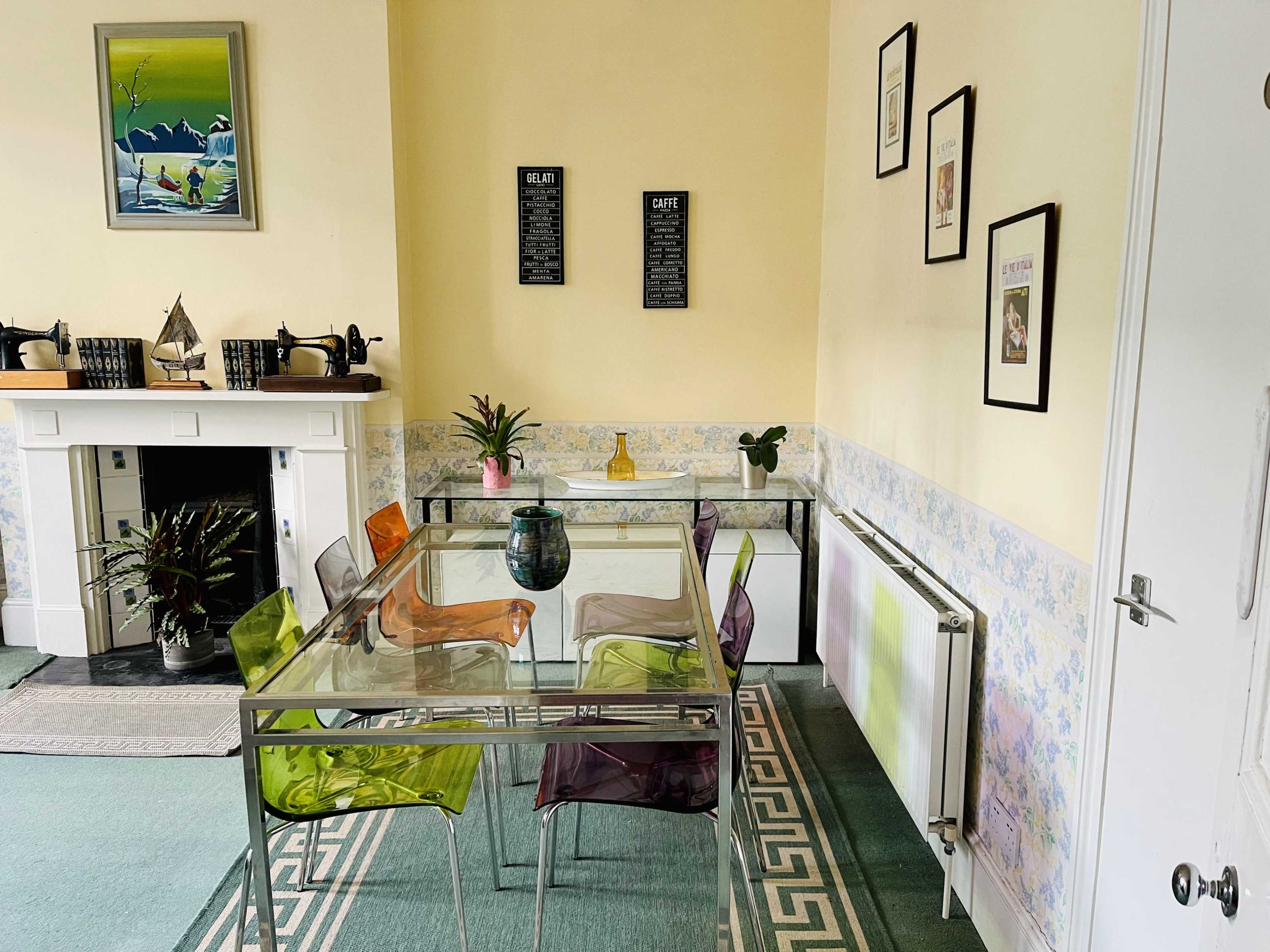 A dining area featuring a glass table surrounded by colorful acrylic chairs, with framed artwork on the walls and a fireplace partially visible in the background.