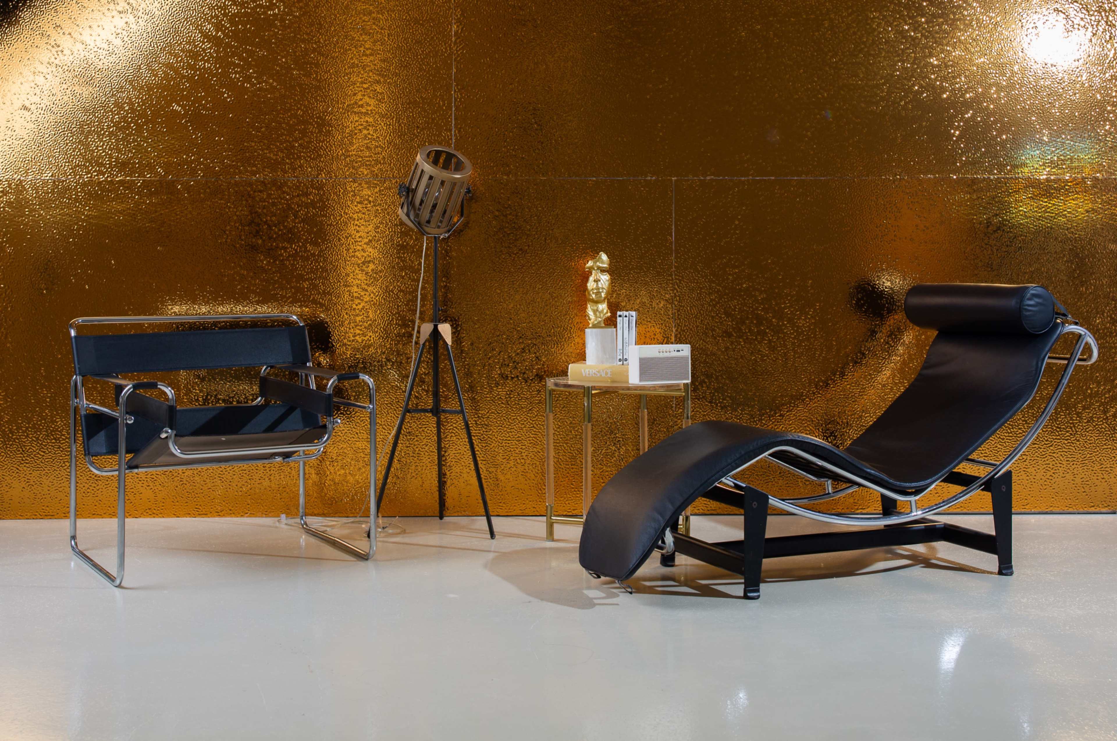 The image features a modern interior setting with a black chaise lounge, a sleek metal chair, a side table displaying a sculpture, and a vintage microphone against a shimmering gold backdrop.