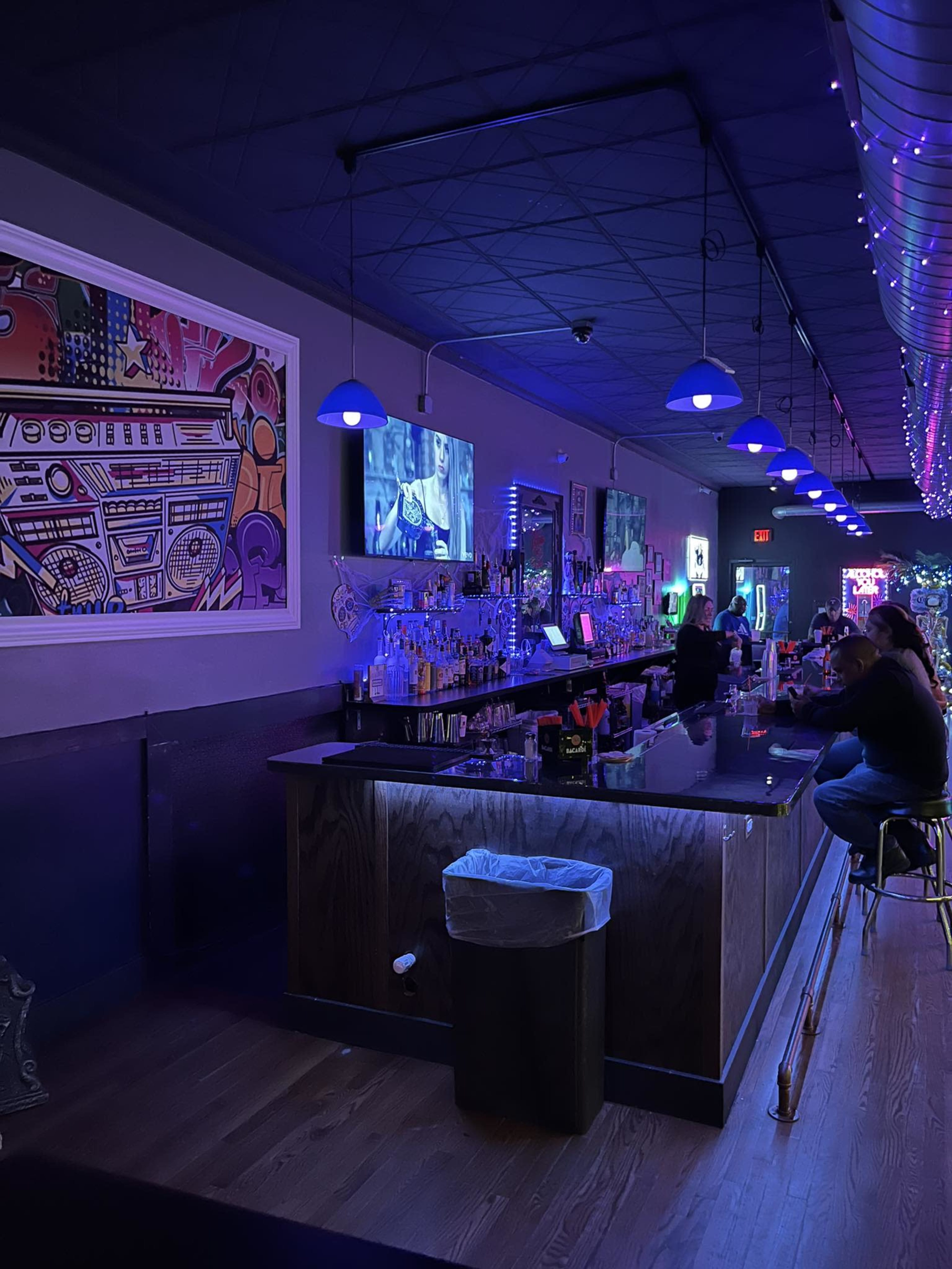 A dimly lit bar with colorful artwork on the walls and a long bar counter where patrons are seated.