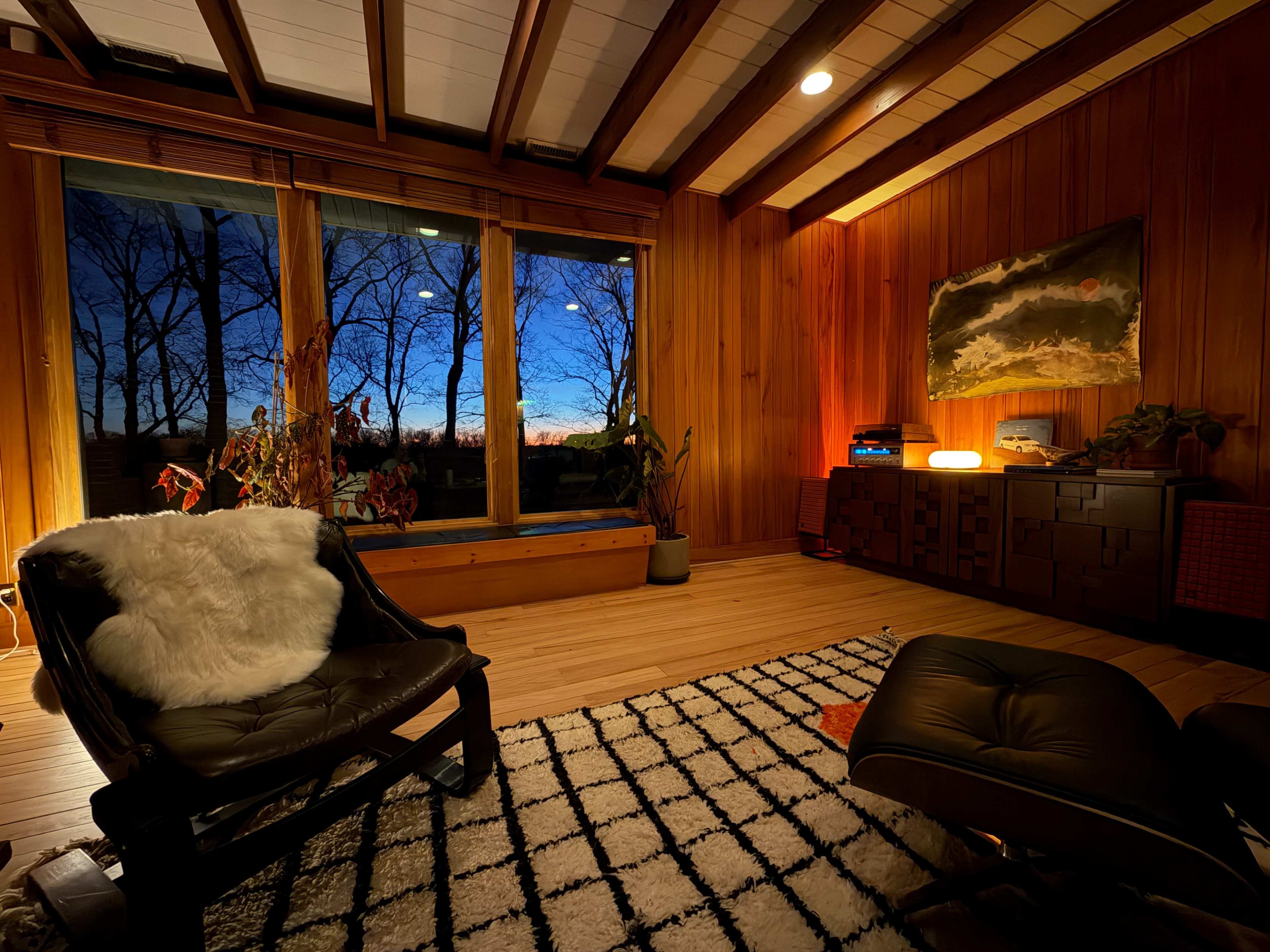 A cozy living room features wooden walls, a large window displaying a sunset, and minimalist furniture, including a black chair and a patterned rug.