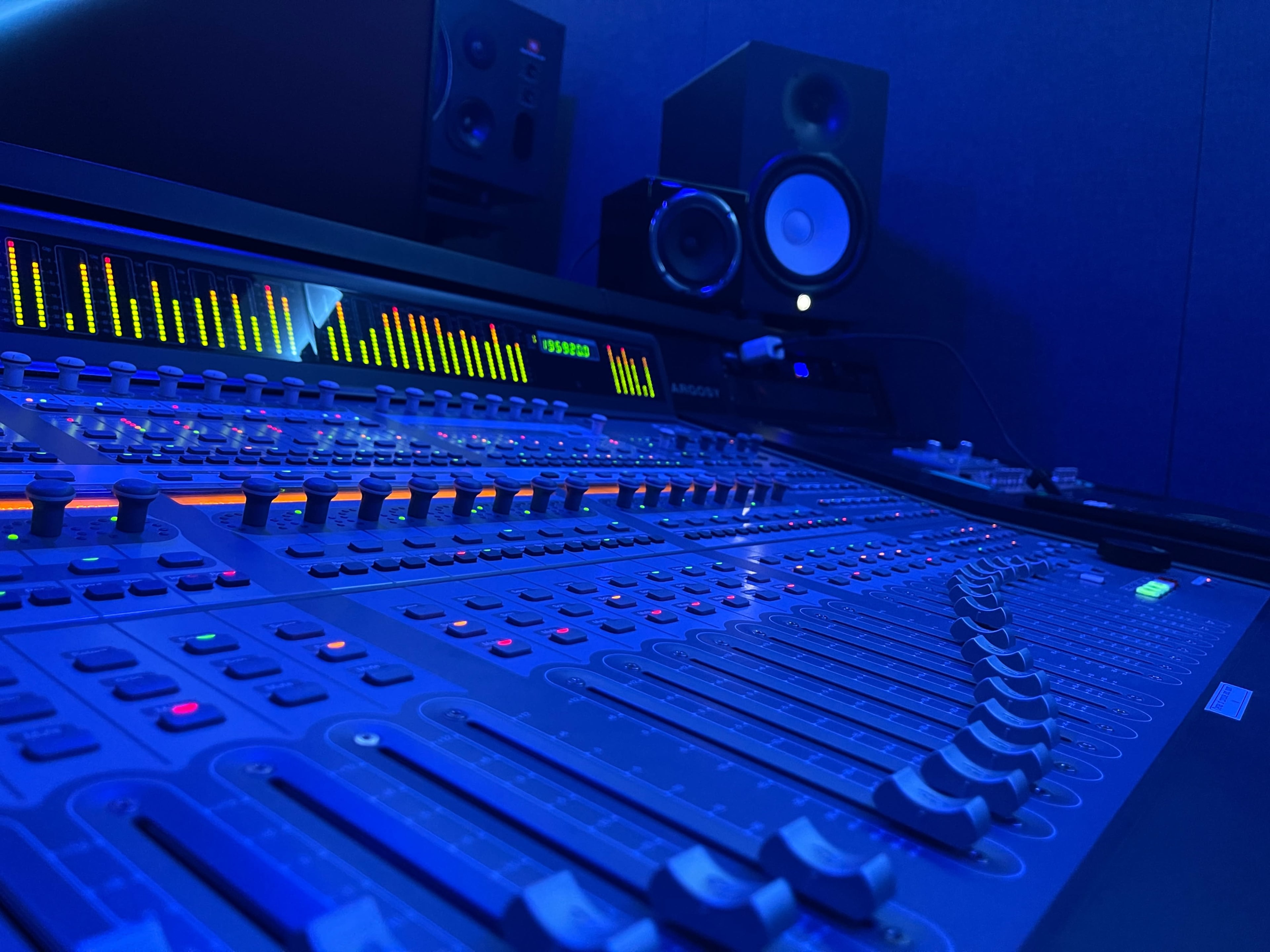 A close-up of a sound mixing console with illuminated controls and speakers in the background.