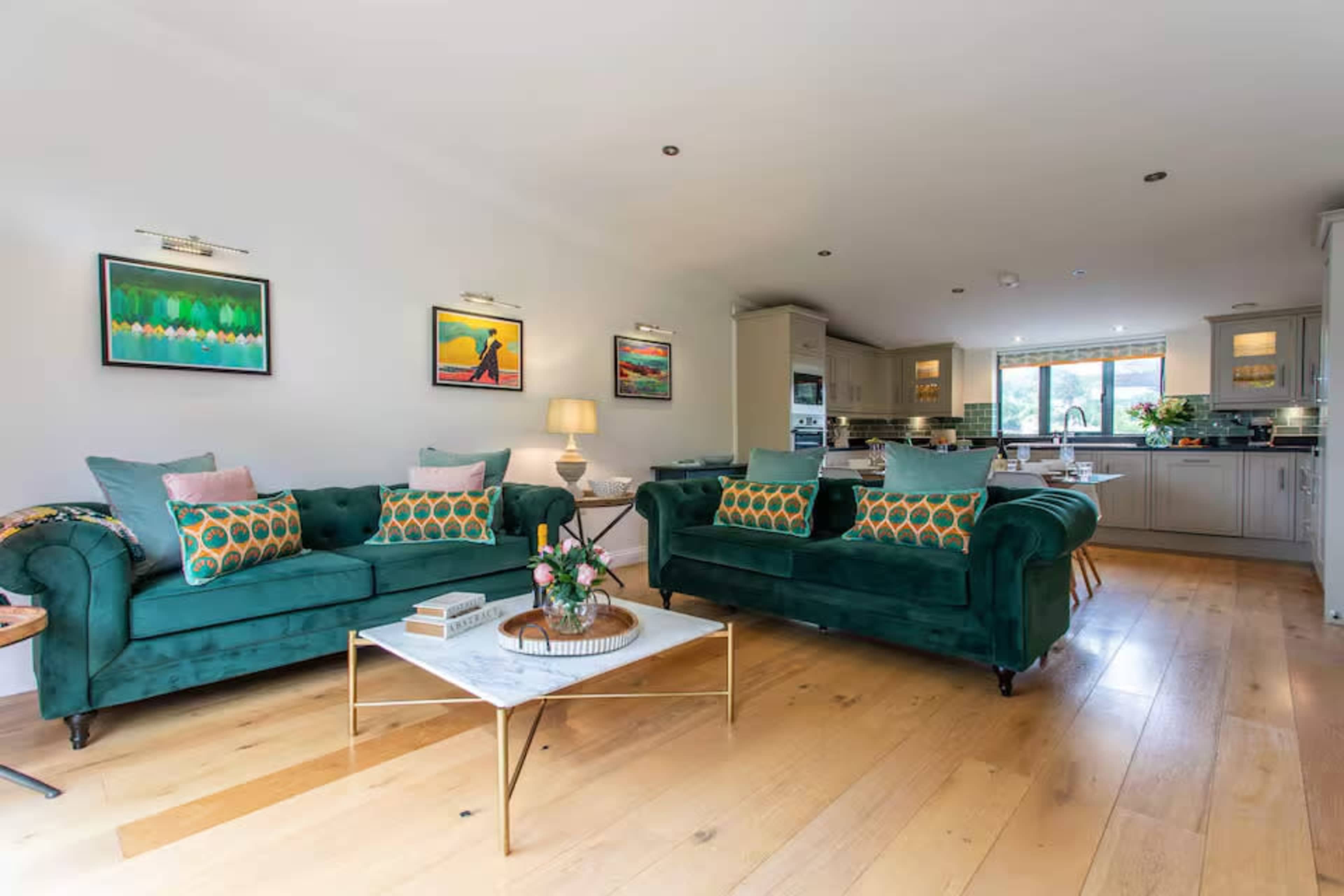 The image shows a modern living room with two green sofas, decorative pillows, and a coffee table, all illuminated by natural light from a nearby window.