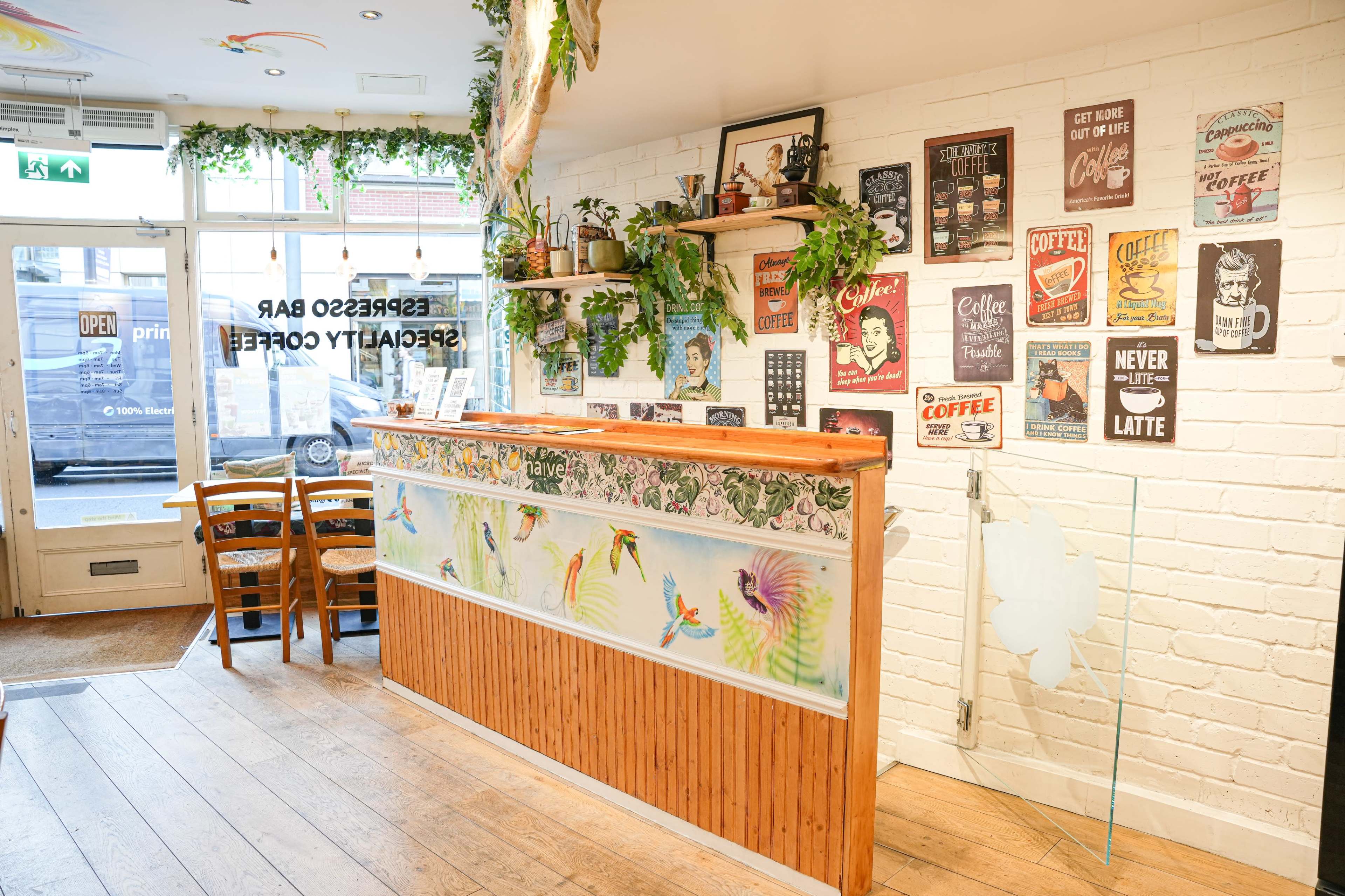 A coffee shop interior with a wooden counter, decorative plant elements, and various coffee-themed posters on the wall.
