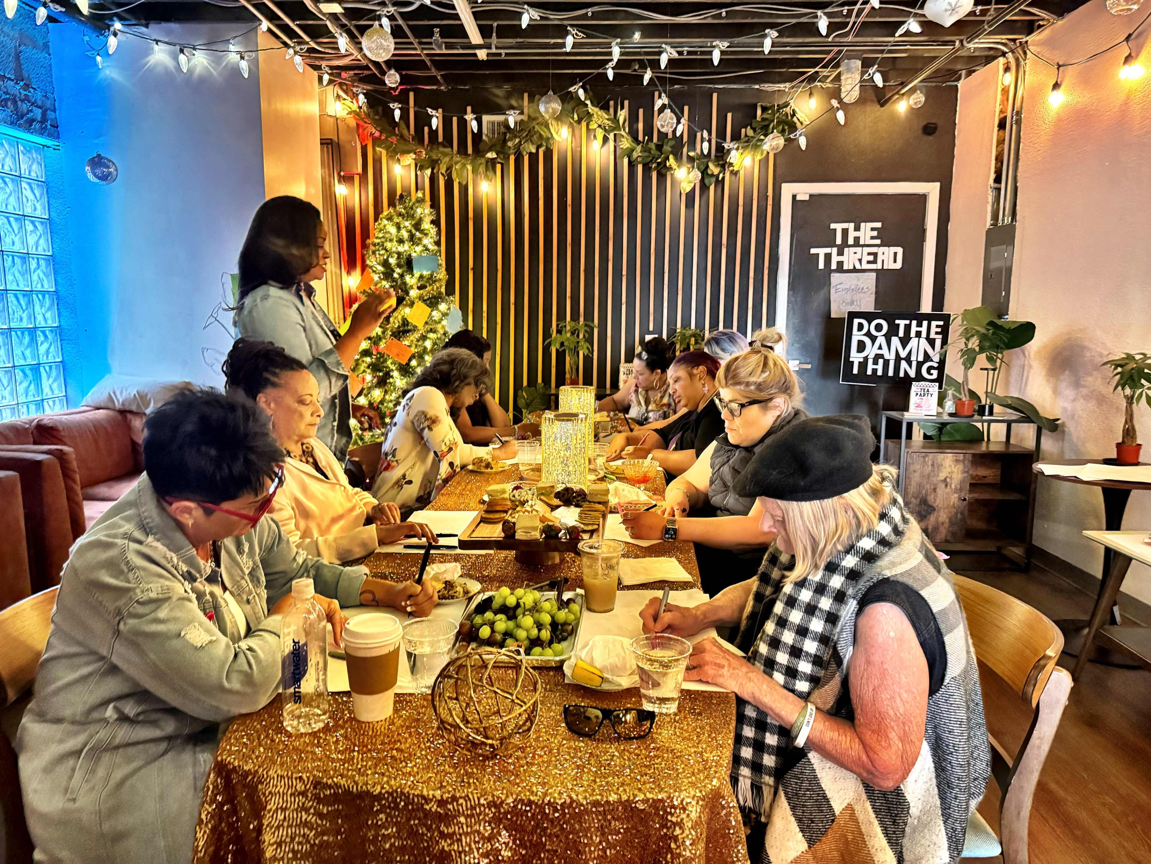A group of people sits around a table adorned with a glittering tablecloth, engaged in conversation and activities in a warmly lit café decorated for the holidays.