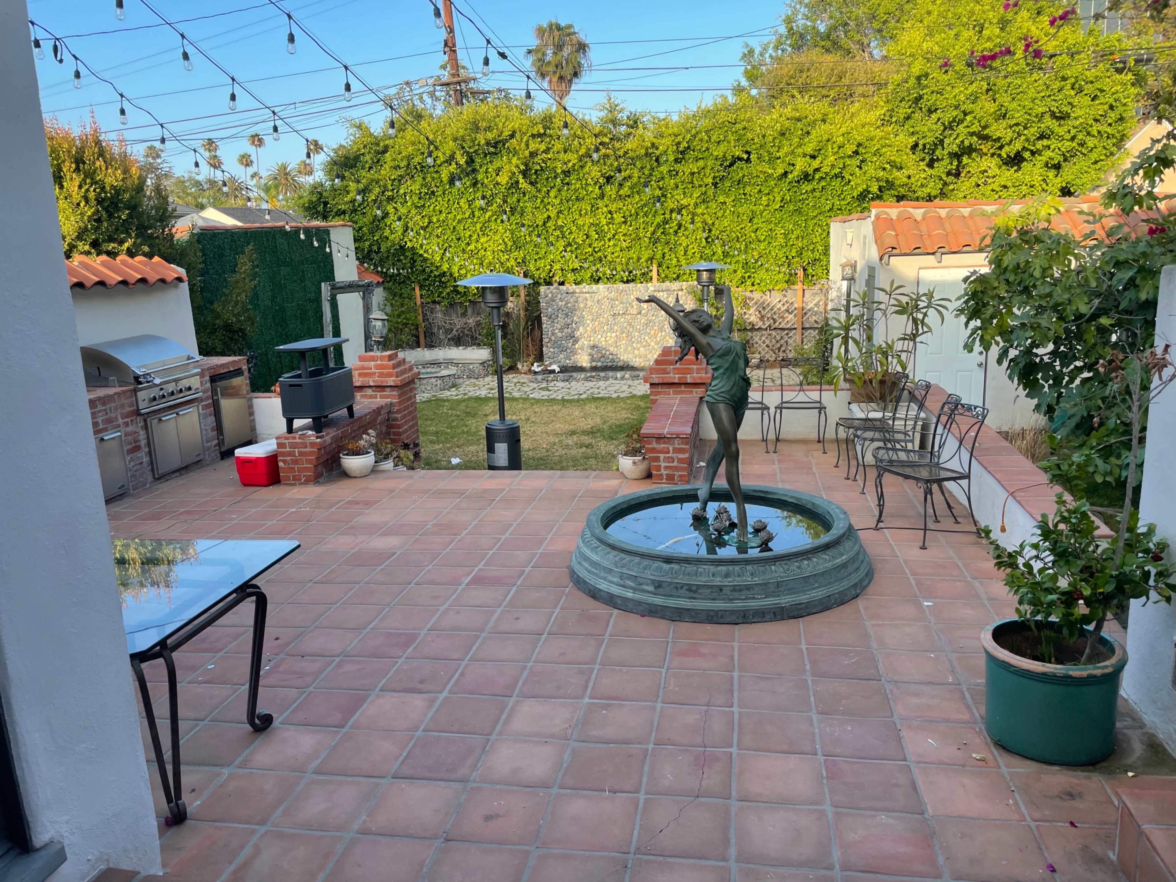 The image shows a outdoor patio area featuring a fountain with a statue, surrounding chairs, a gas grill, and string lights overhead.