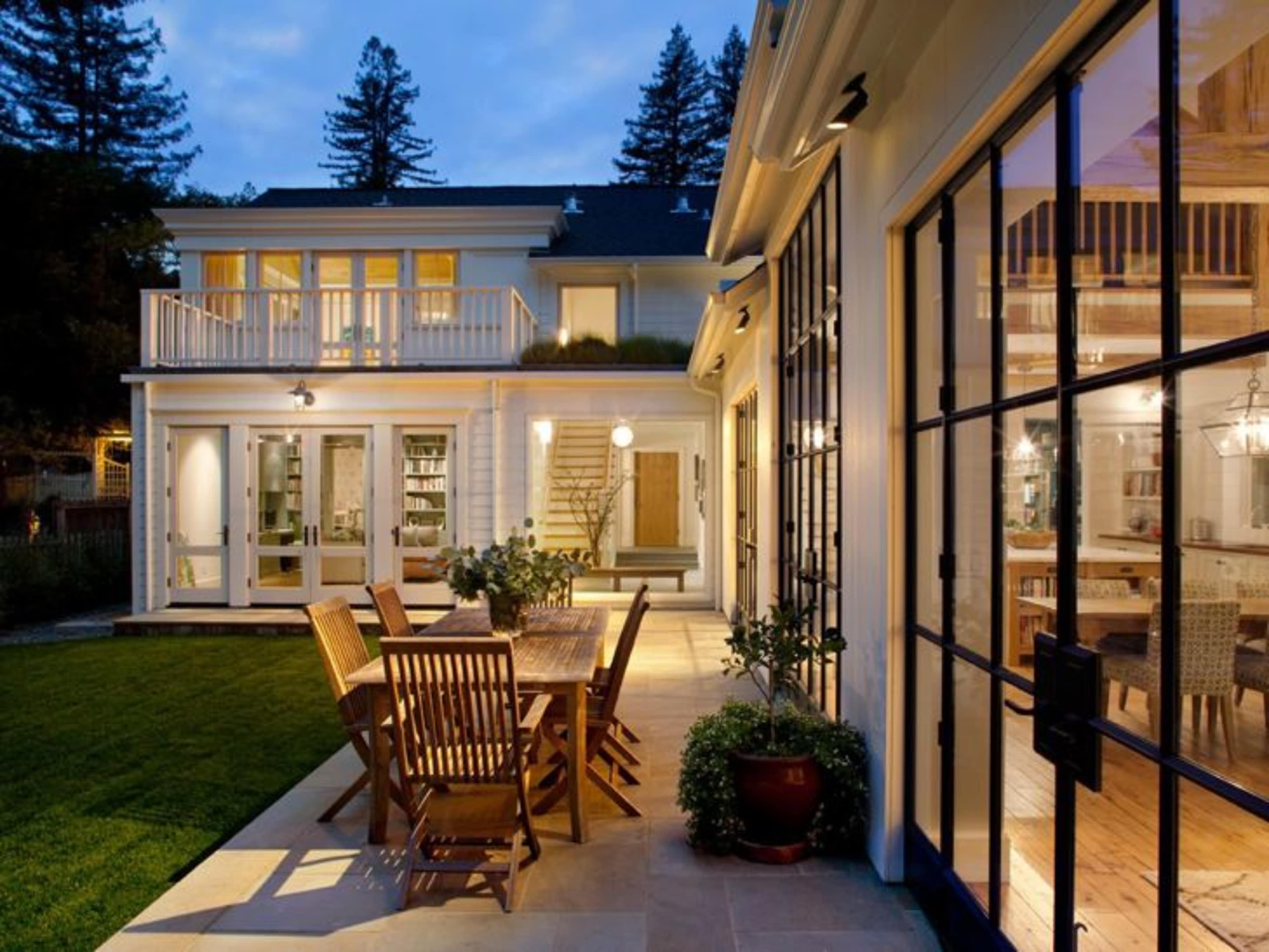 A modern two-story house with large glass doors, a patio dining set, and a landscaped garden illuminated by outdoor lighting at dusk.